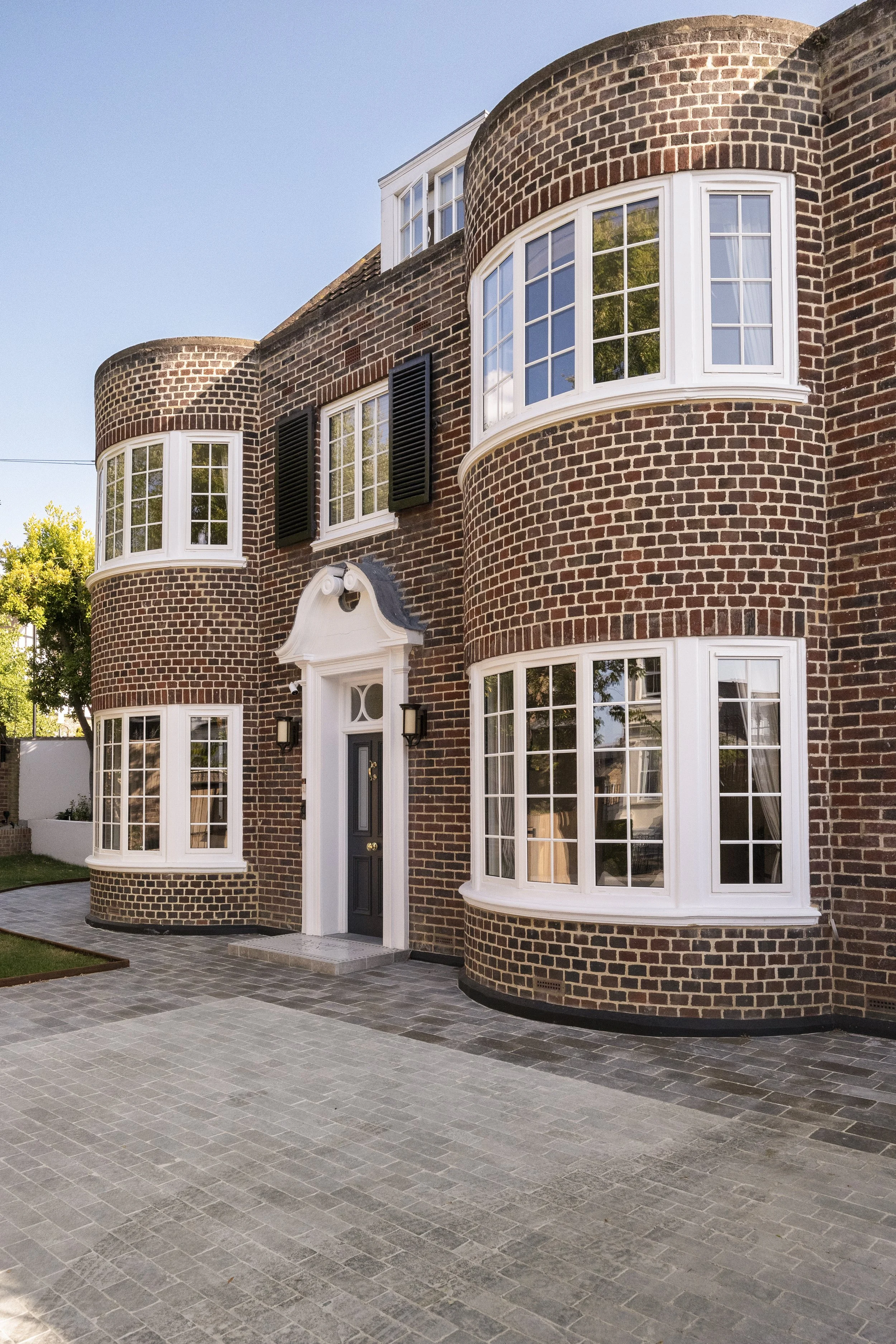 A brick residential building with multiple large white-framed windows, black shutters, a black front door, and a small covered porch with black lamps on either side. The building is surrounded by a paved courtyard with gray bricks and some greenery v