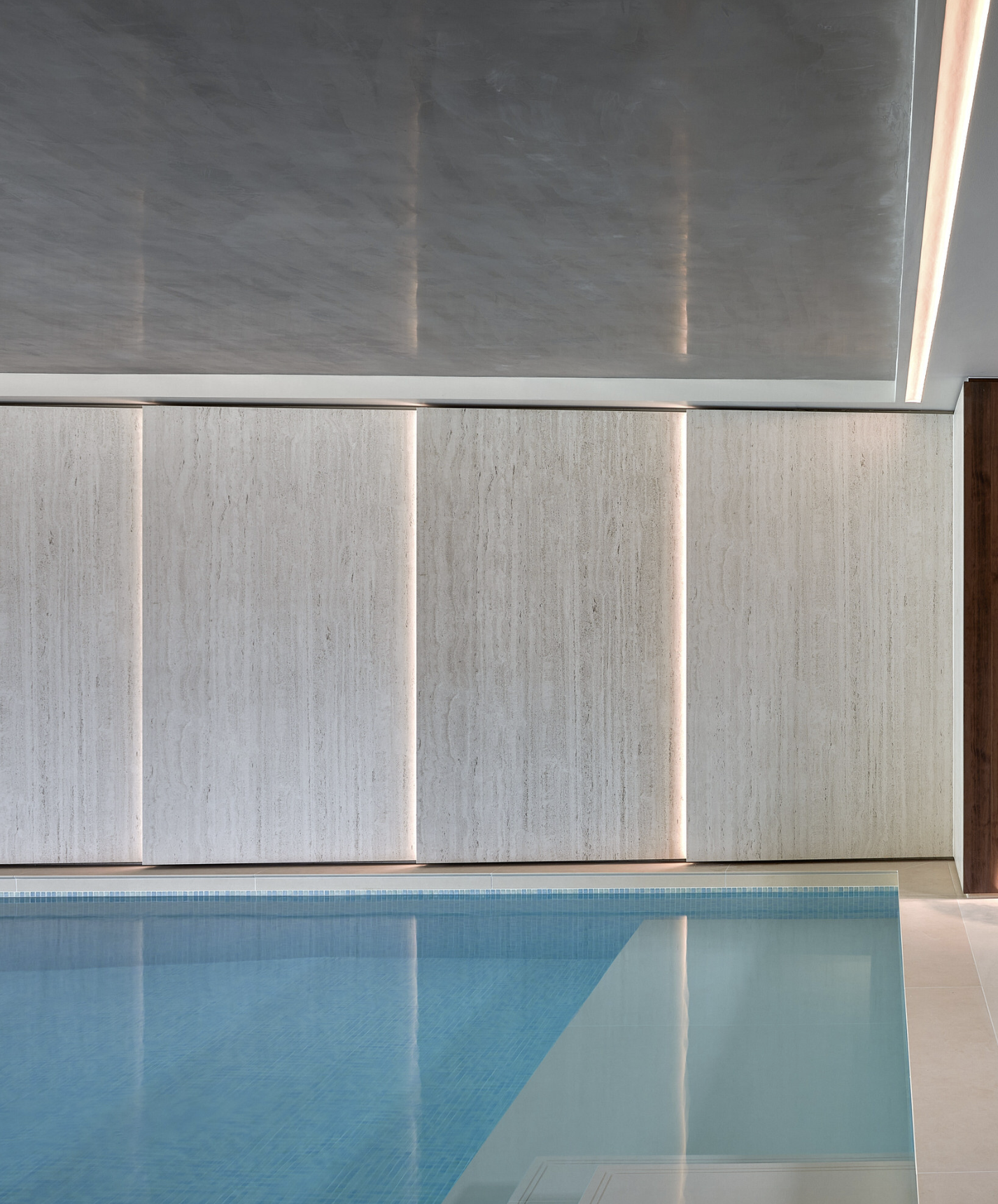 Indoor swimming pool with a reflection on the water, neutral-toned walls with vertical light strips, and a ceiling with recessed lighting.