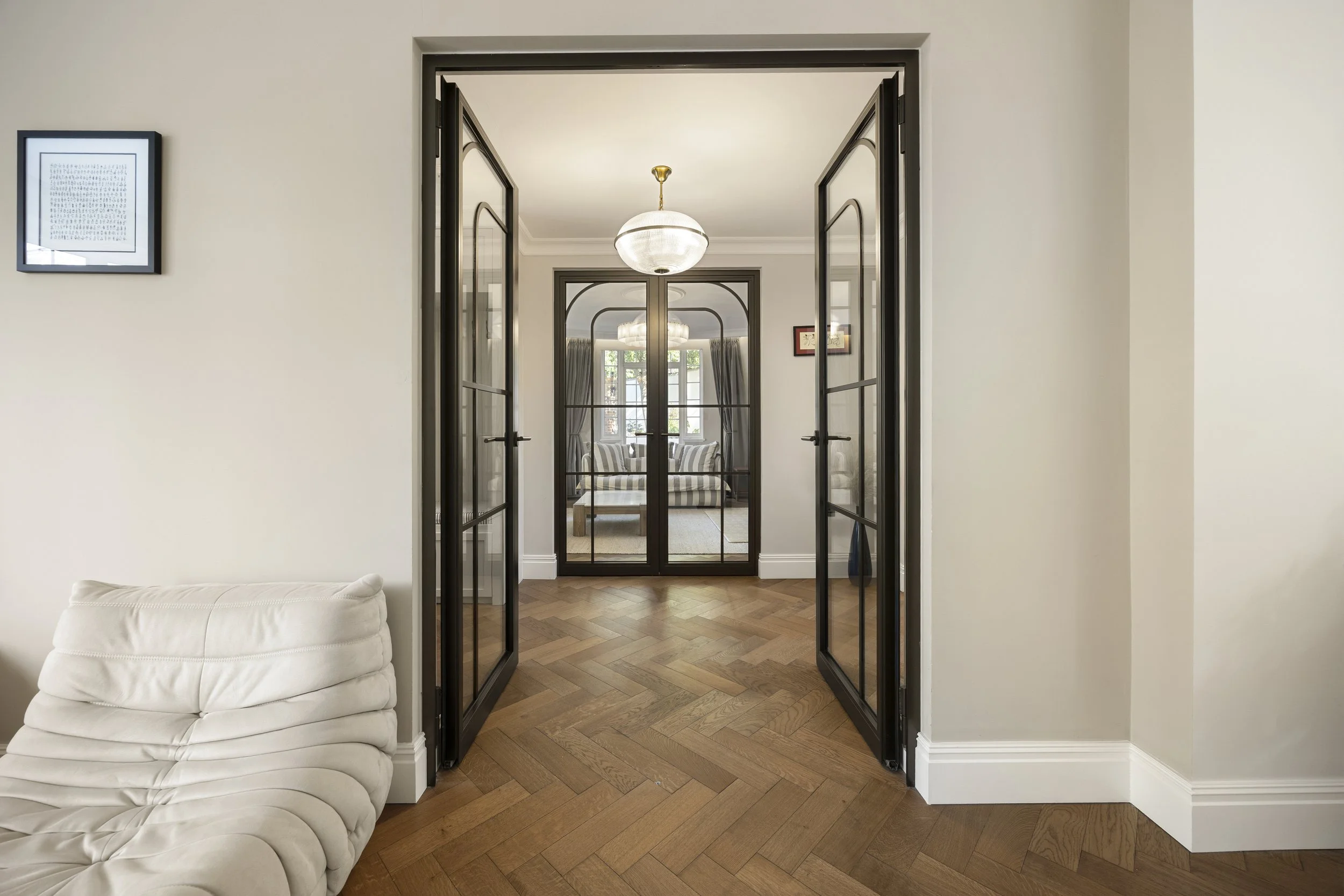 A view through black-framed glass double doors into a living room with a white couch, patterned curtains, and a chandelier, with light-colored walls and hardwood floors.
