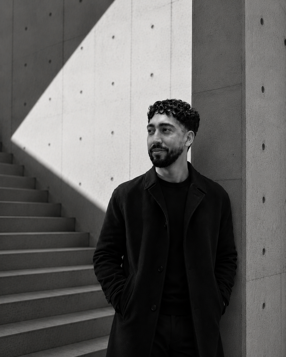 A man with curly hair and a beard wearing a black coat looks to the side while standing outdoors near concrete stairs and modern concrete wall with a geometric shadow.