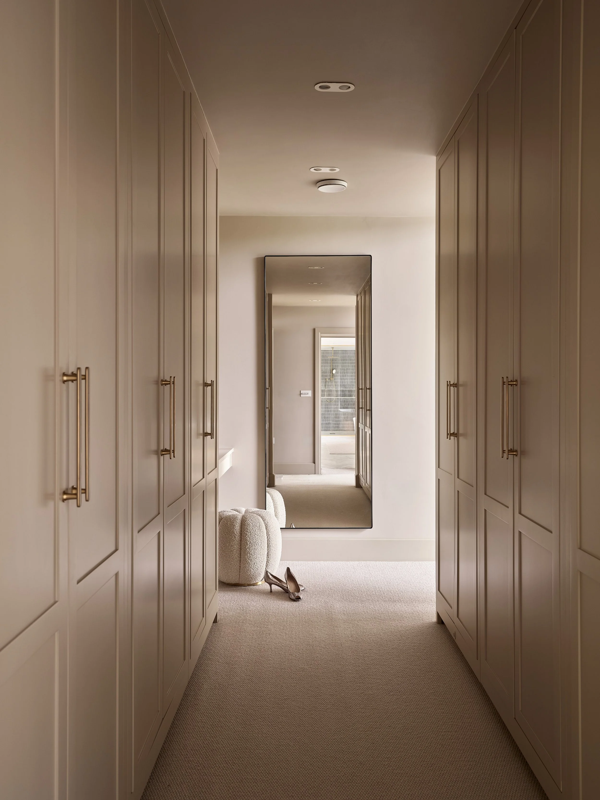 A hallway with built-in closets on both sides, a large mirror on the right wall, a plush ottoman near the mirror, and a pair of high-heeled shoes on the floor. Doorways lead to other rooms with natural light coming through an outdoor area.