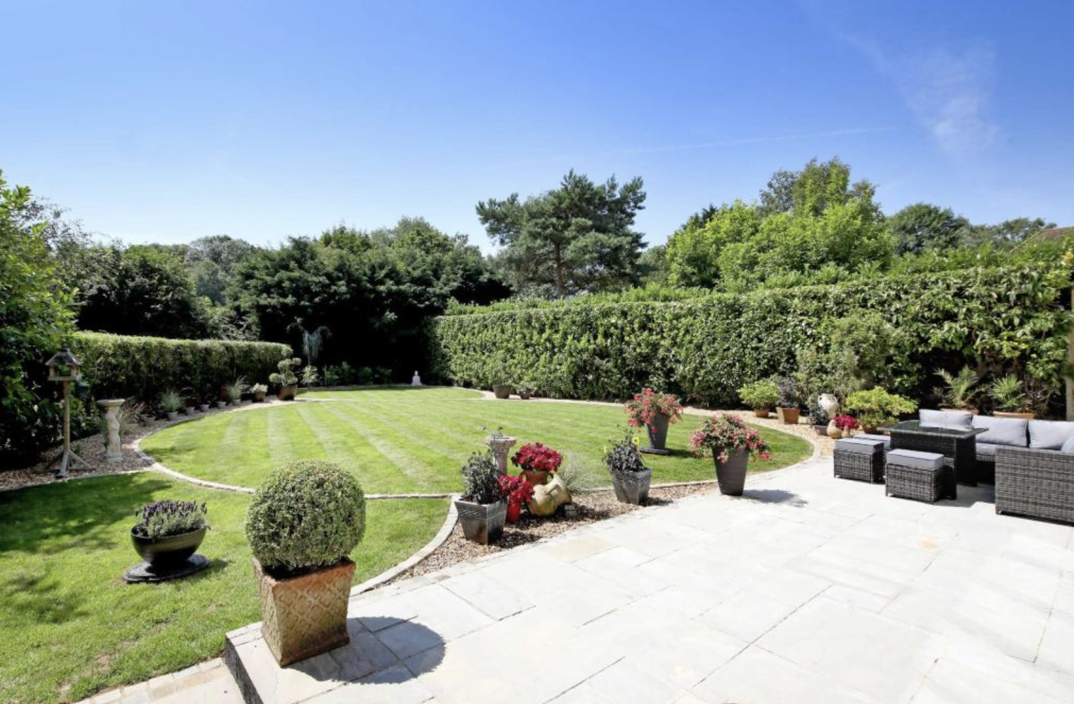 A backyard garden with a manicured lawn, potted flowers, hedges, and outdoor seating under a blue sky.