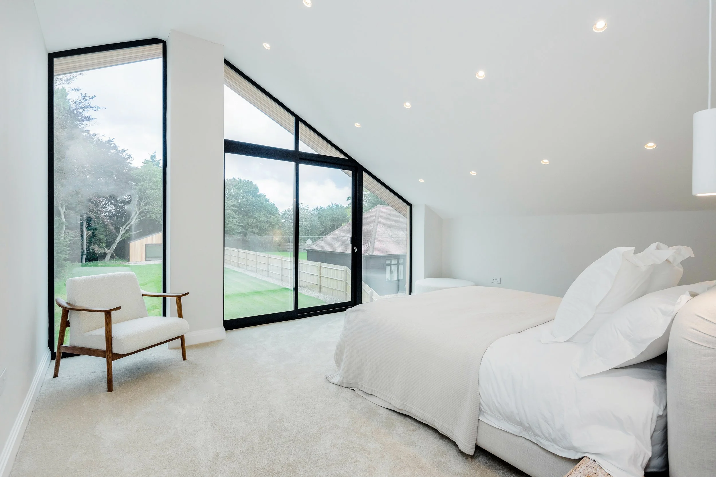 Modern bedroom with large floor-to-ceiling windows, white bedding, beige carpet, and a white chair with wooden arms