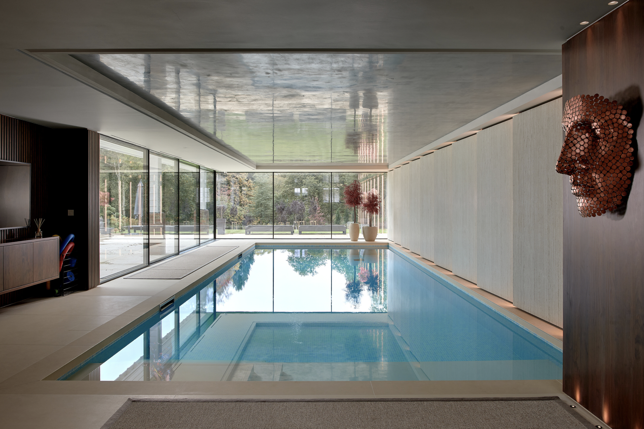 Indoor swimming pool with large windows showing trees outside, modern decor, and a red sculpture of a human face on the wall.