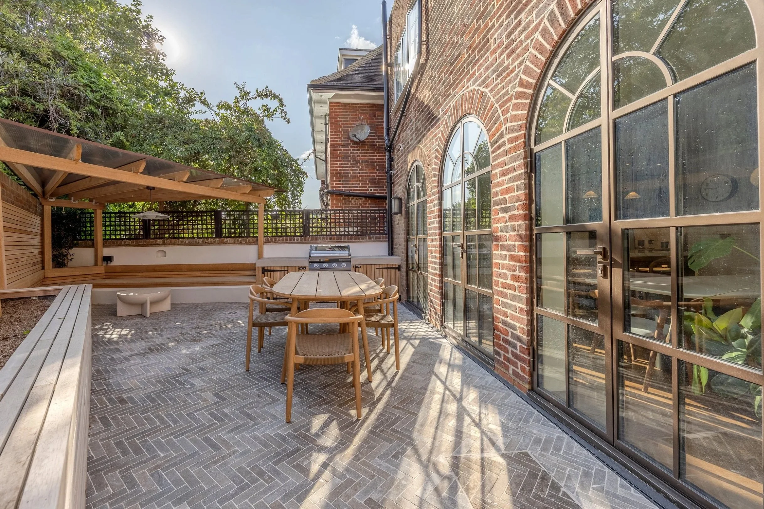 A backyard patio with a round wooden dining table and six matching chairs. A barbecue grill is situated against the brick wall of the house. The patio has a decorative gray brick floor and is partially covered by a wooden pergola. There is a black wo