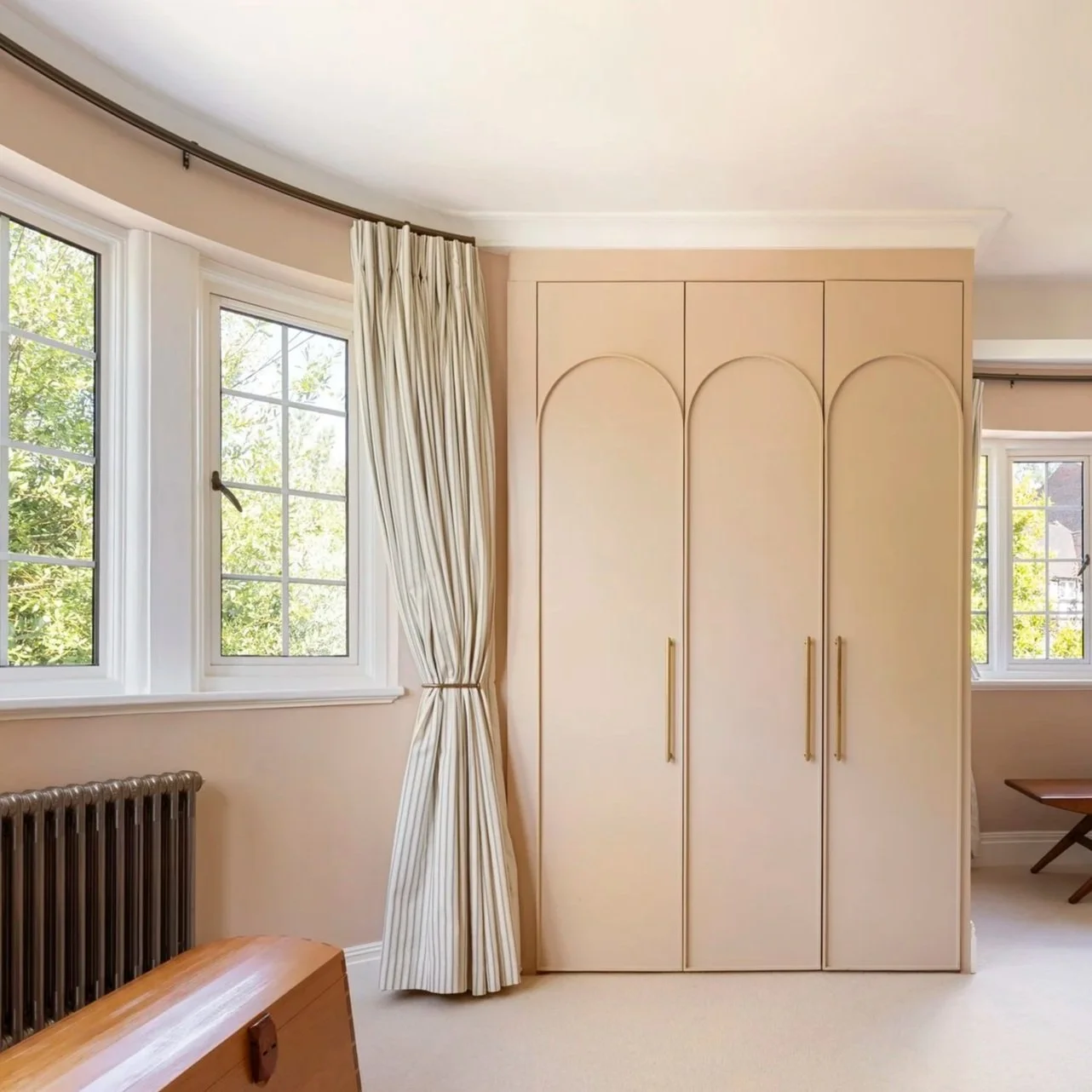 Interior view of a room with two windows, cream-colored curtains, a radiator, and a built-in closet with gold handles and arched detailing. There is a wooden chest in the foreground.