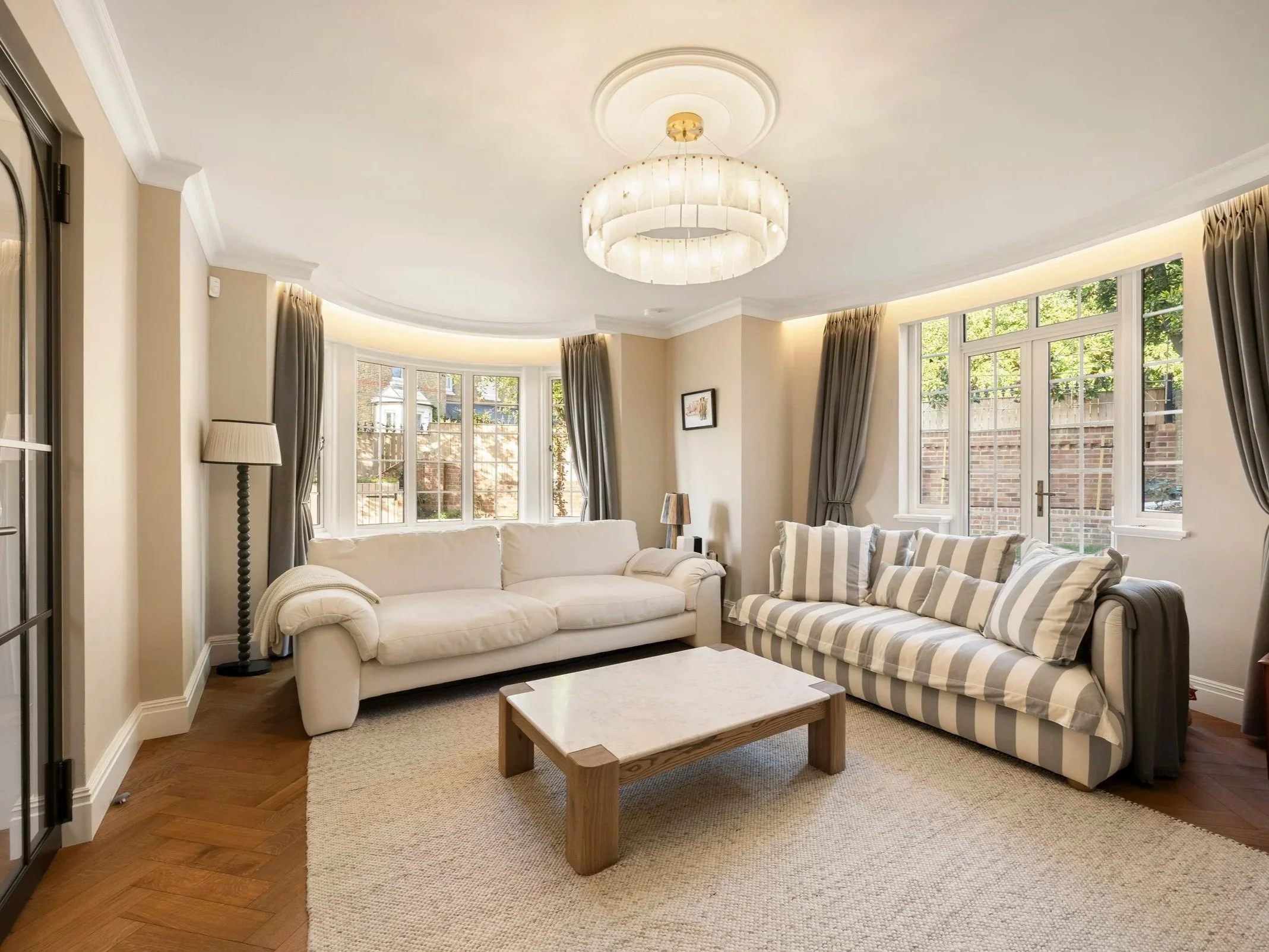Living room with beige walls, large windows with brown curtains, white and striped sofas, a wooden coffee table, a floor lamp, and a ceiling light fixture.