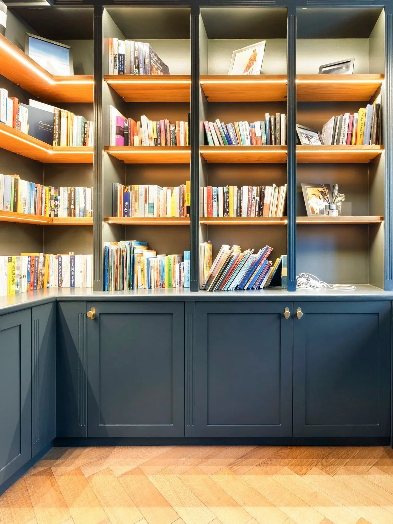 Dark blue bookshelf with wooden shelves filled with books, framed photos, and small decorative items, situated above dark blue cabinets with gold knobs on a wooden floor.