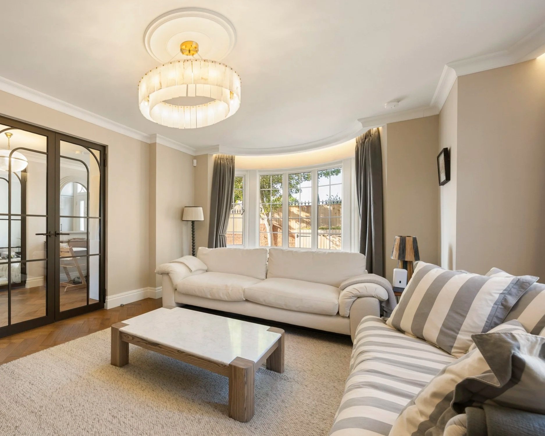 Living room with cream-colored walls, a white sofa, striped armchair, coffee table, large bay window with curtains, and a ceiling light fixture.
