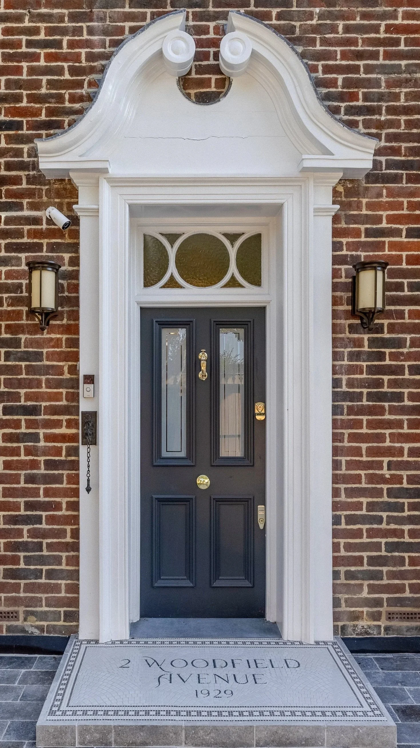 Front door with black paint, rectangular panels, brass hardware, and a transom window with decorative circles, set in a brick wall with two wall-mounted lamps and a welcome mat.