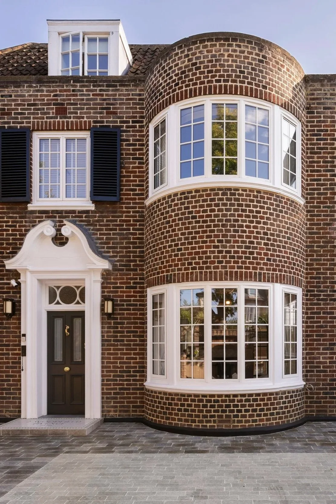 Front view of a traditional brick house with black front door, white trim, and large bay windows with white frames. The house has a decorative white door surround with lanterns on either side.