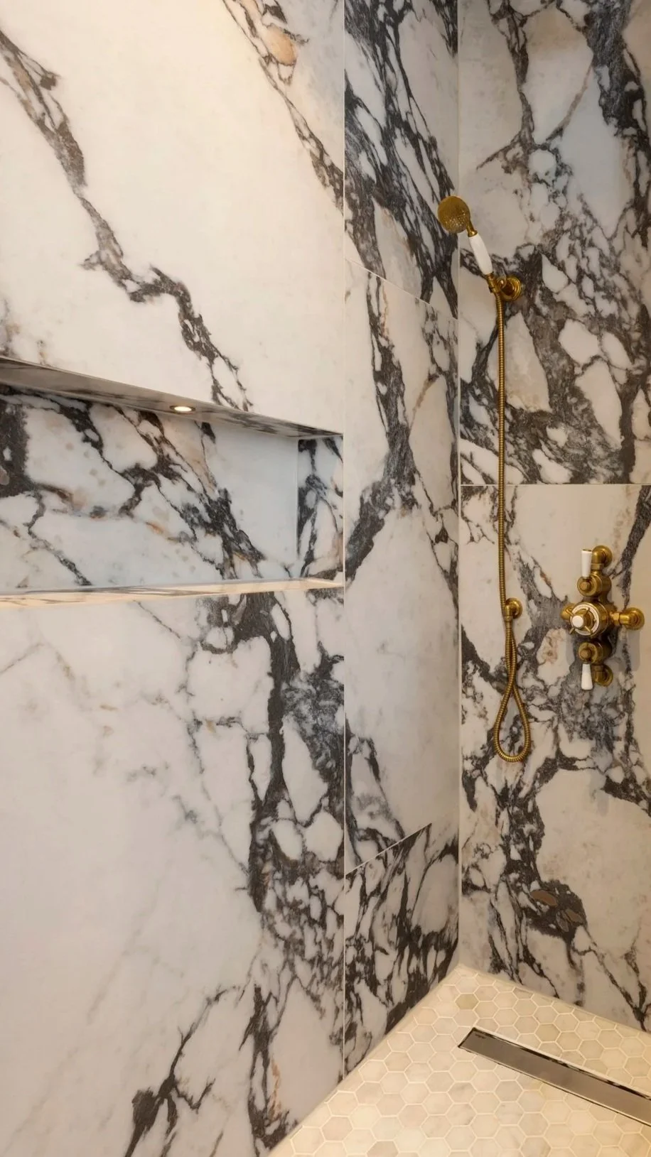 Marble shower walls with gold fixtures, including a handheld showerhead and control knobs, and a floor with small hexagonal tiles and a drain.