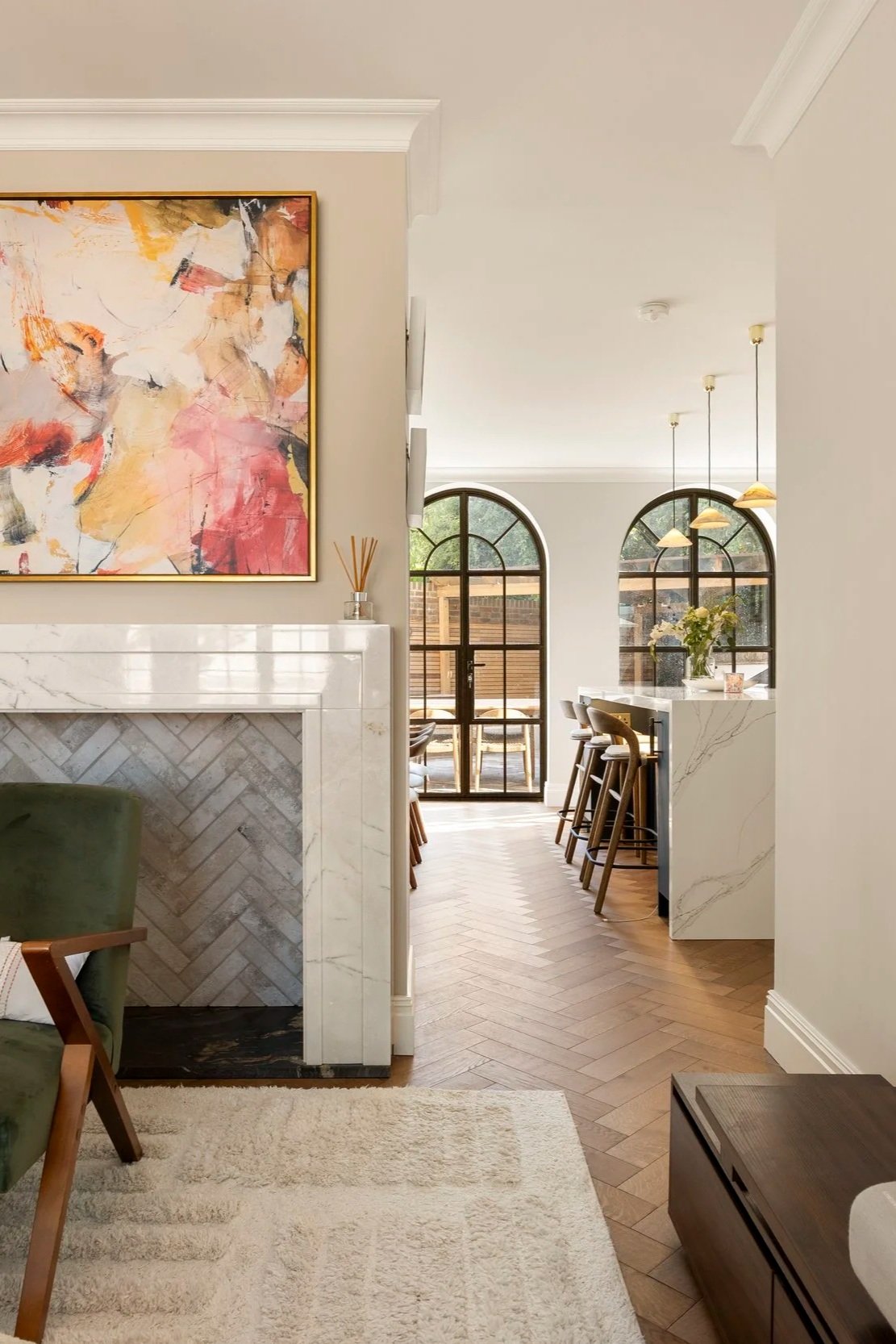 Modern interior view of a home with a white fireplace, marble accents, artwork, and a kitchen area with arched windows, bar stools, and hanging lights.