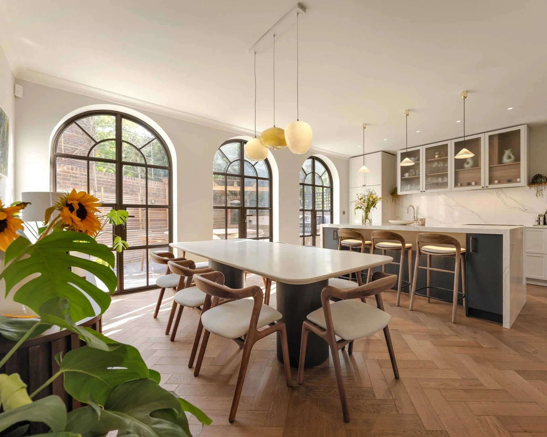 Bright, modern kitchen with large arched windows, white cabinetry, marble backsplash, kitchen island with bar stools, pendant lights, and potted plants.