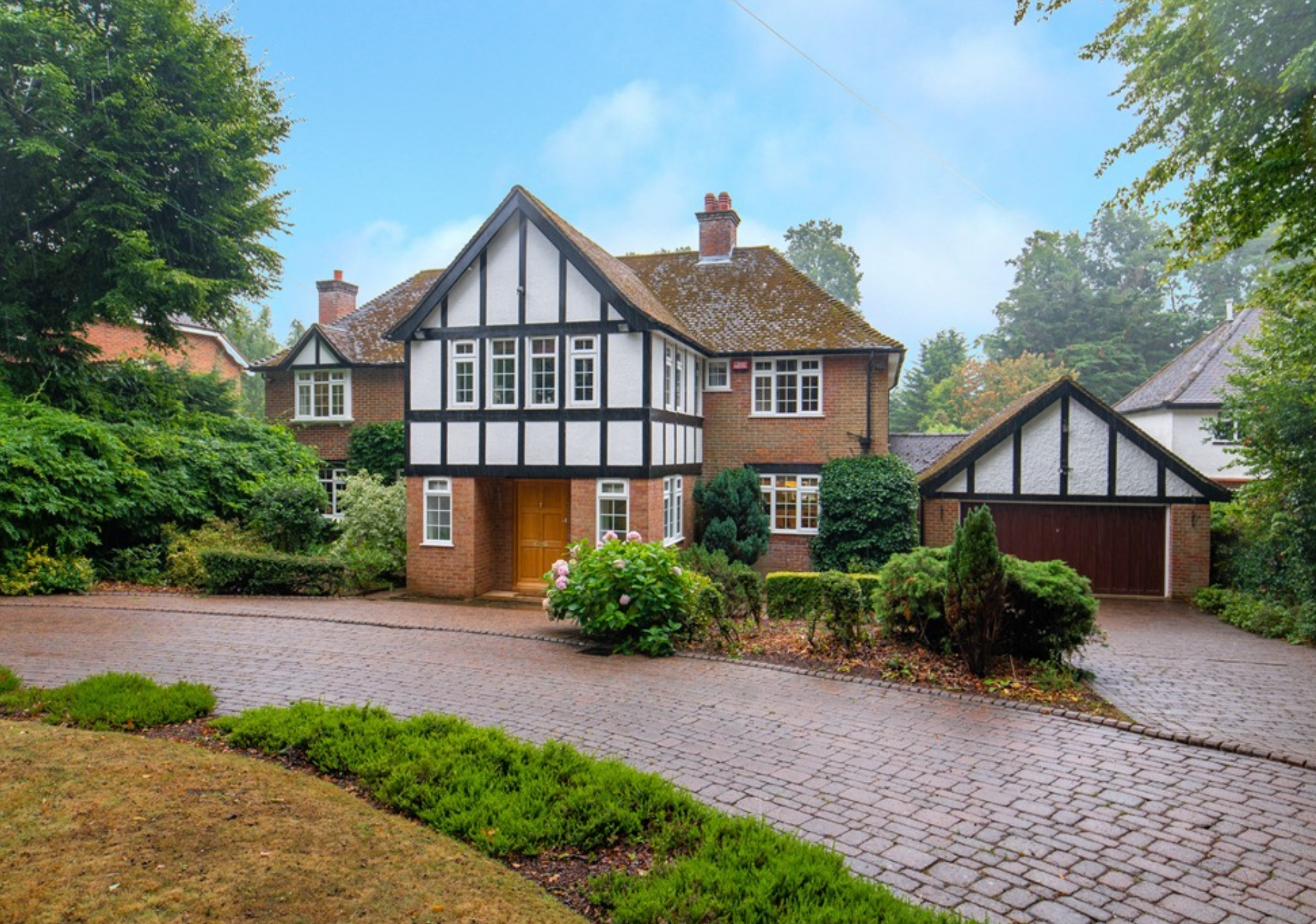 A large house with a black and white Tudor-style facade, red brick walls, and a tiled roof, surrounded by trees and a paved driveway.
