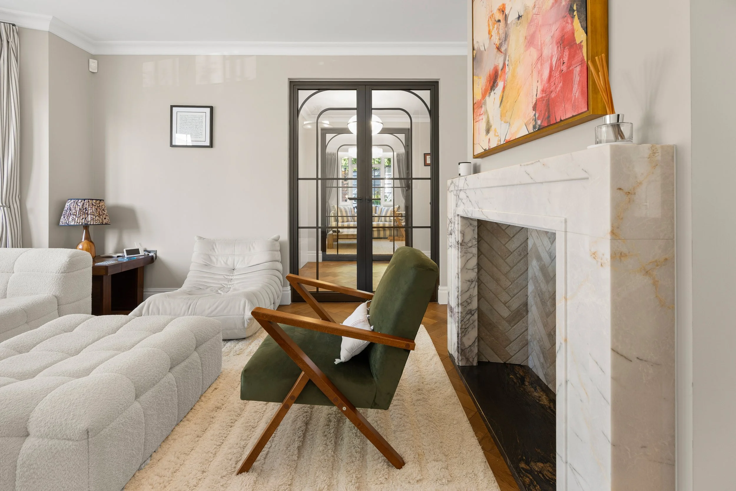 Living room with white and green chairs, a fireplace with marble surround, a large abstract painting, and a doorway with black-framed glass doors leading to a sunlit room with striped furniture.