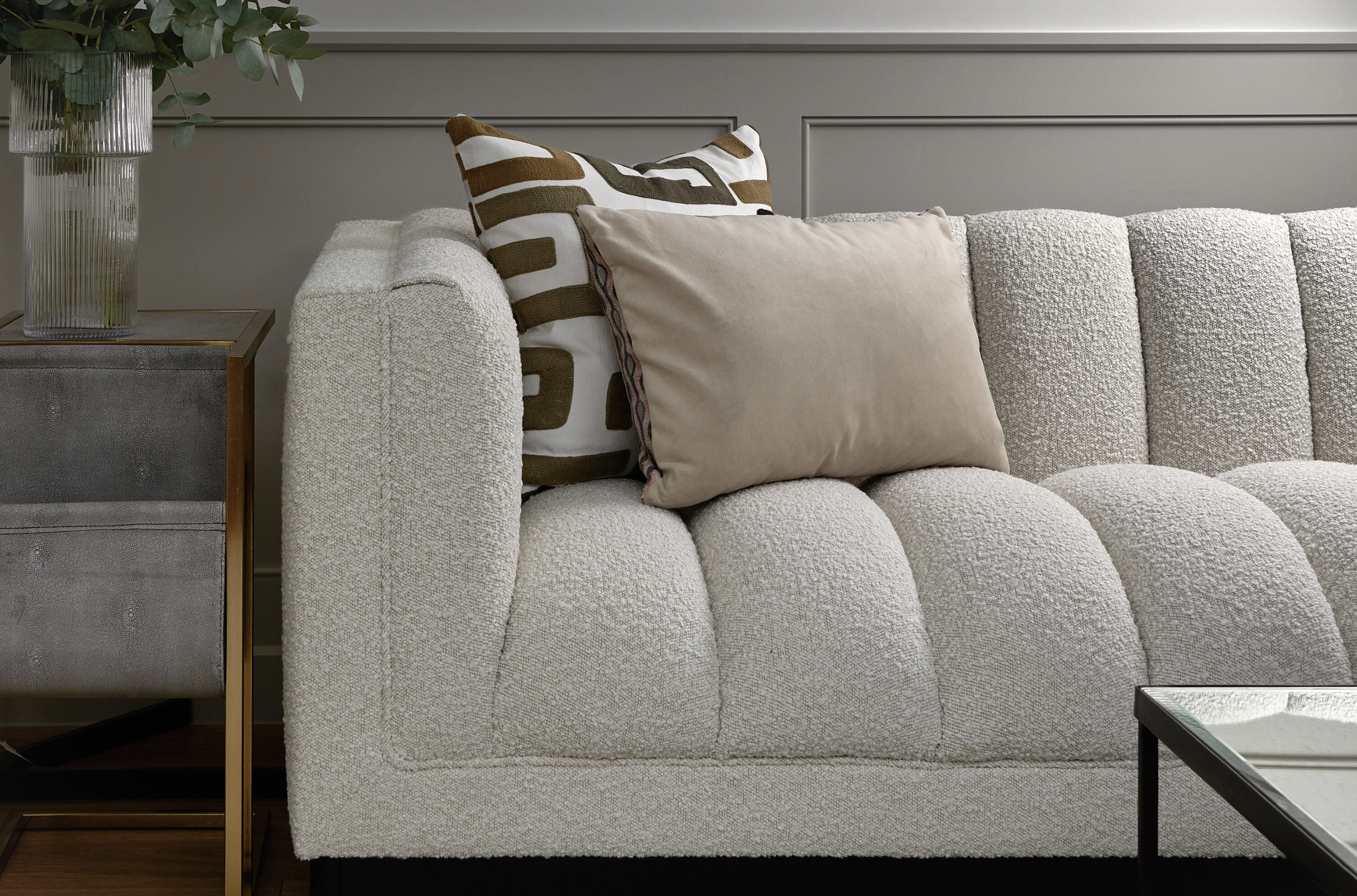 A beige textured sofa with two decorative pillows, one with a patterned design and the other plain, placed against a gray wall. A side table with a glass vase of green foliage is on the left, and a black metal table is partially visible on the right.