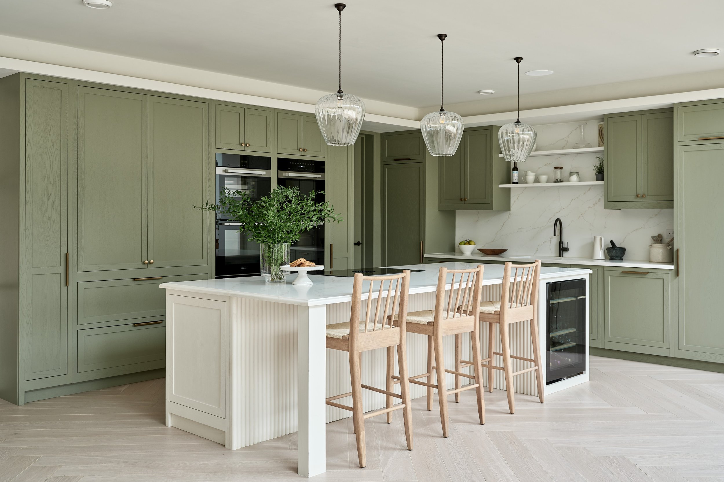 Modern kitchen with sage green cabinets, a white island with beadboard detailing, and three glass pendant lights over the island. There are wooden bar stools at the island and an open shelf with white containers and decorative items.