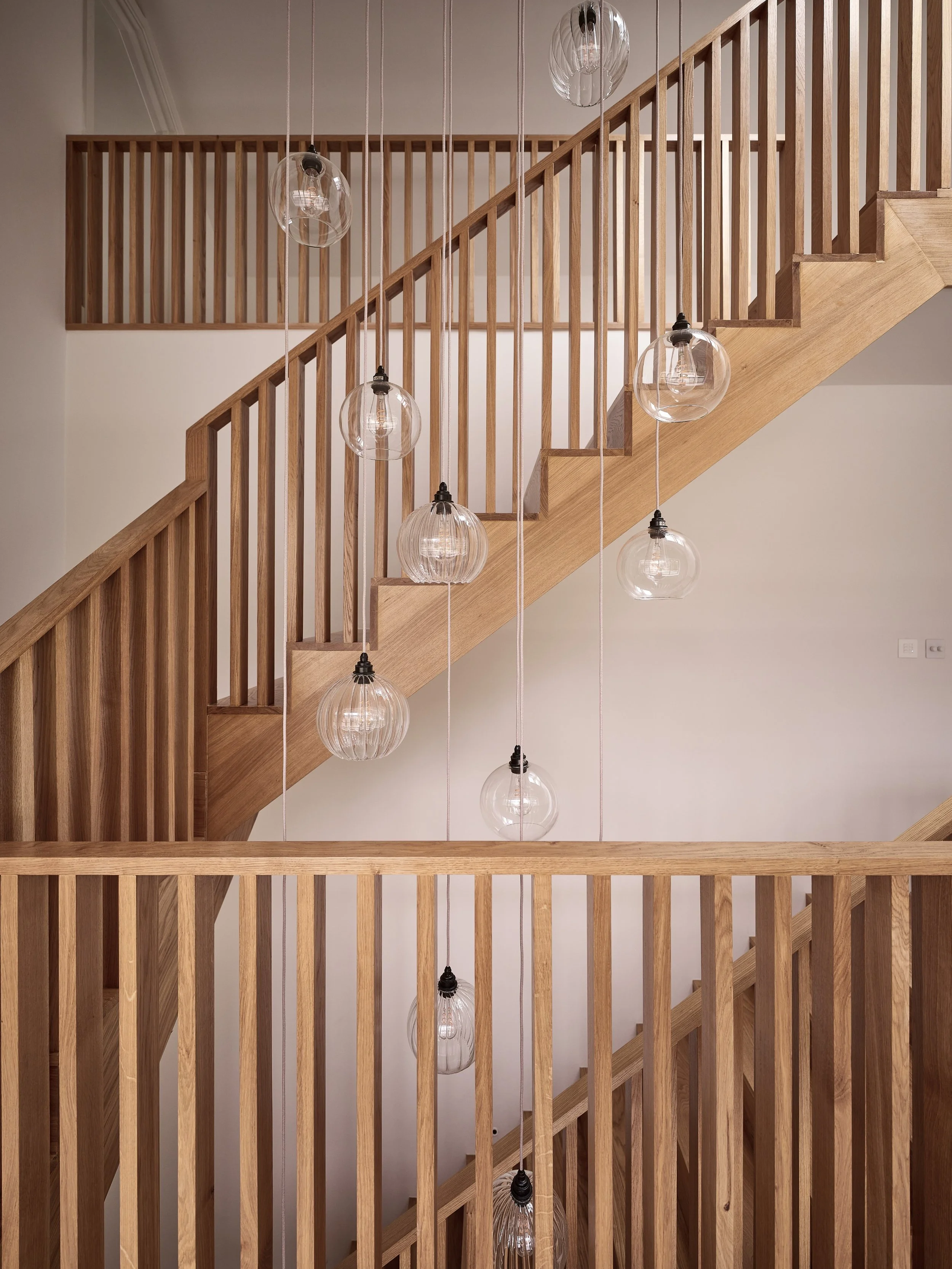 Wooden staircase with vertical railing, hanging glass pendant lights, and a second-floor wooden railing in the background.