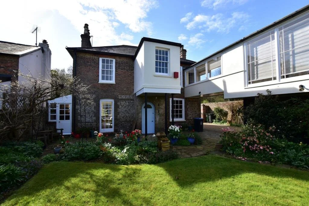 A backyard view of a two-story brick house with white-framed windows and a blue front door, surrounded by a well-maintained lawn, flower beds with blooming plants, and a stone pathway, with a neighboring house and a covered porch on the right.