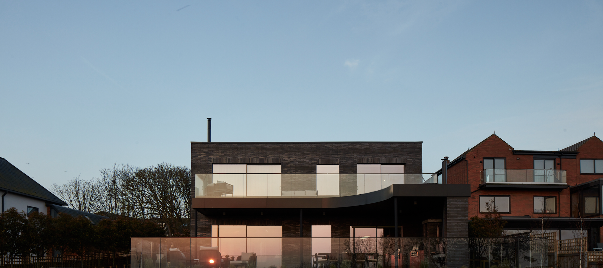 Modern brick house with large glass windows and balcony, set against a clear blue sky with surrounding trees.