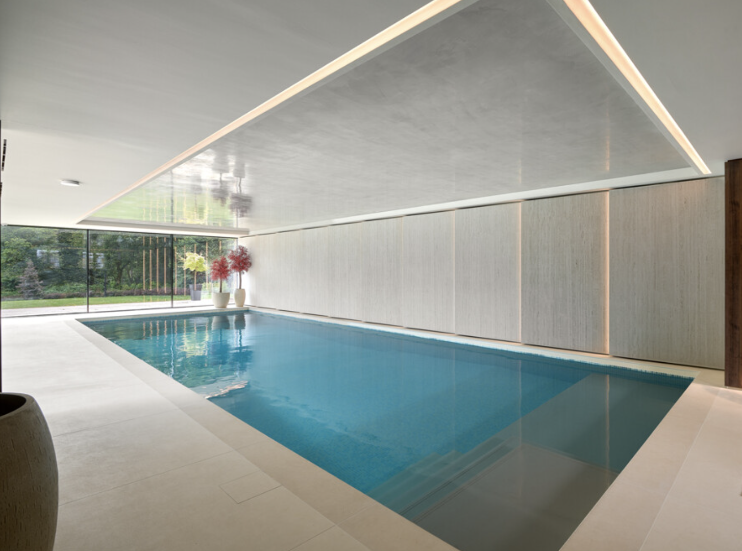 Indoor swimming pool with large glass windows showing trees outside, minimalistic decor with potted plants, and a ceiling with built-in lighting.