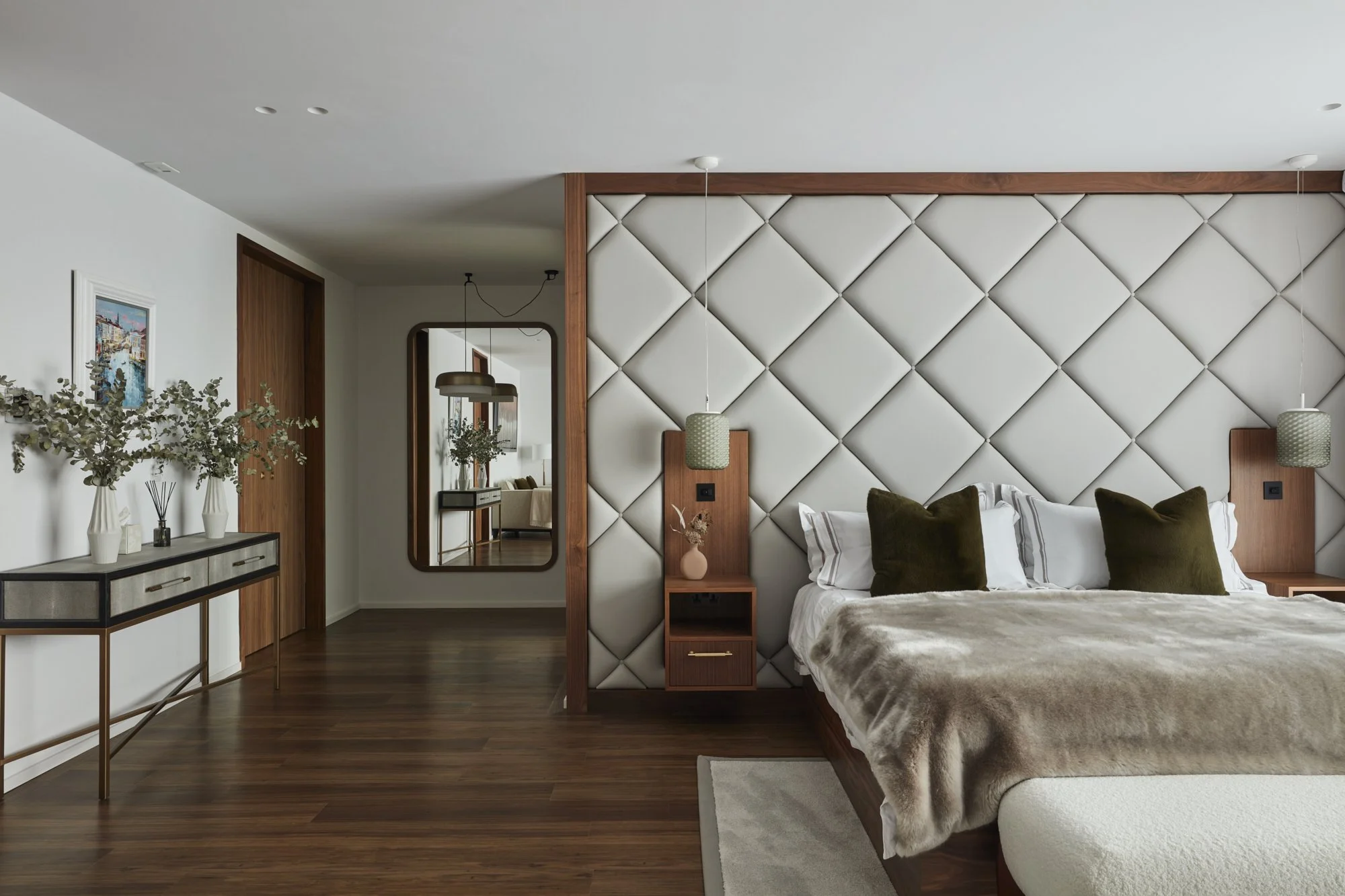Modern bedroom with a bed, nightstand, decorative pillows, and wall-mounted lamps. Full-length mirror, wooden featured wall, and artwork on the wall. Dining area visible through doorway.