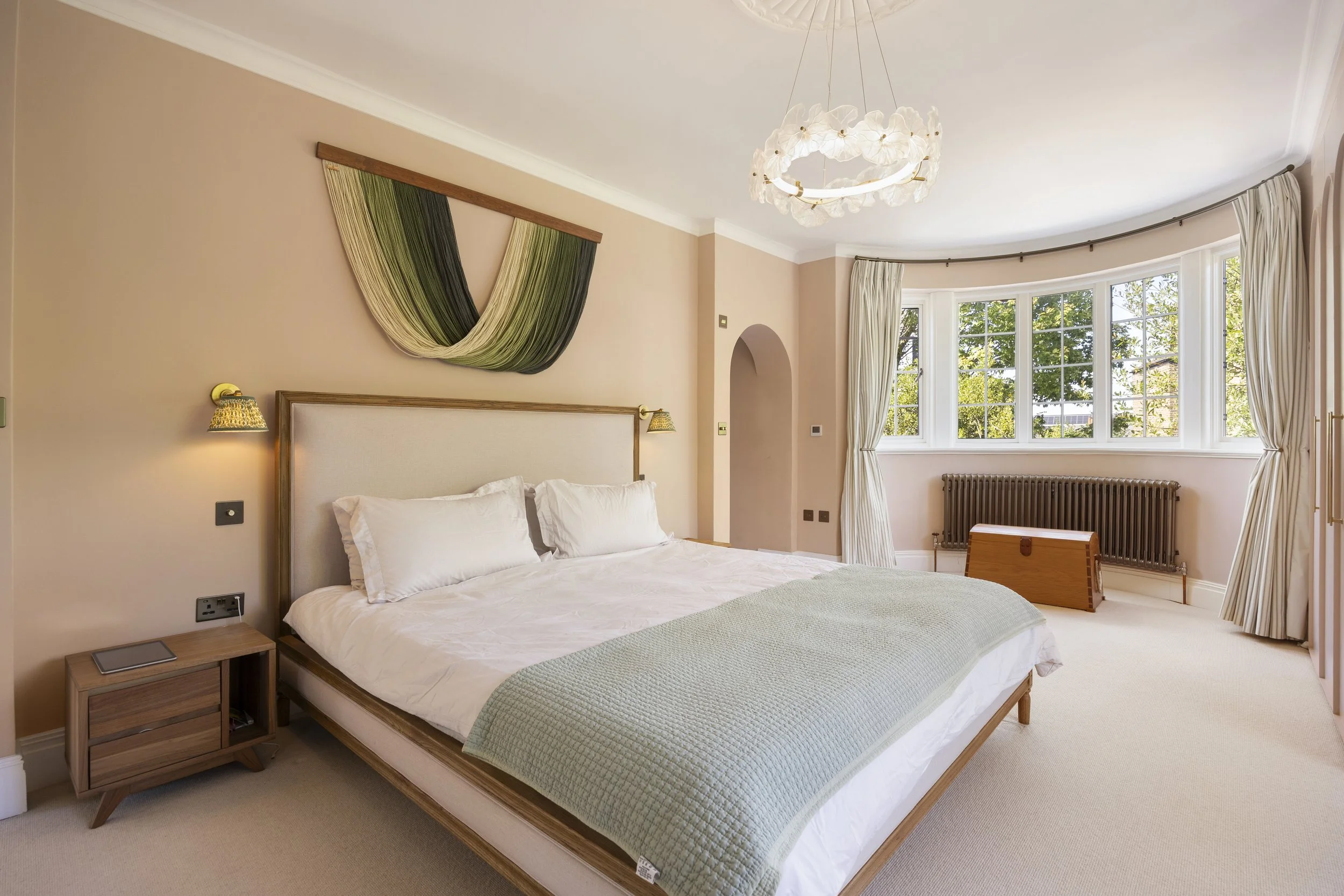 A bedroom with a bed, nightstand, and a large bay window with curtains, a decorative wall hanging, and a ceiling light fixture.