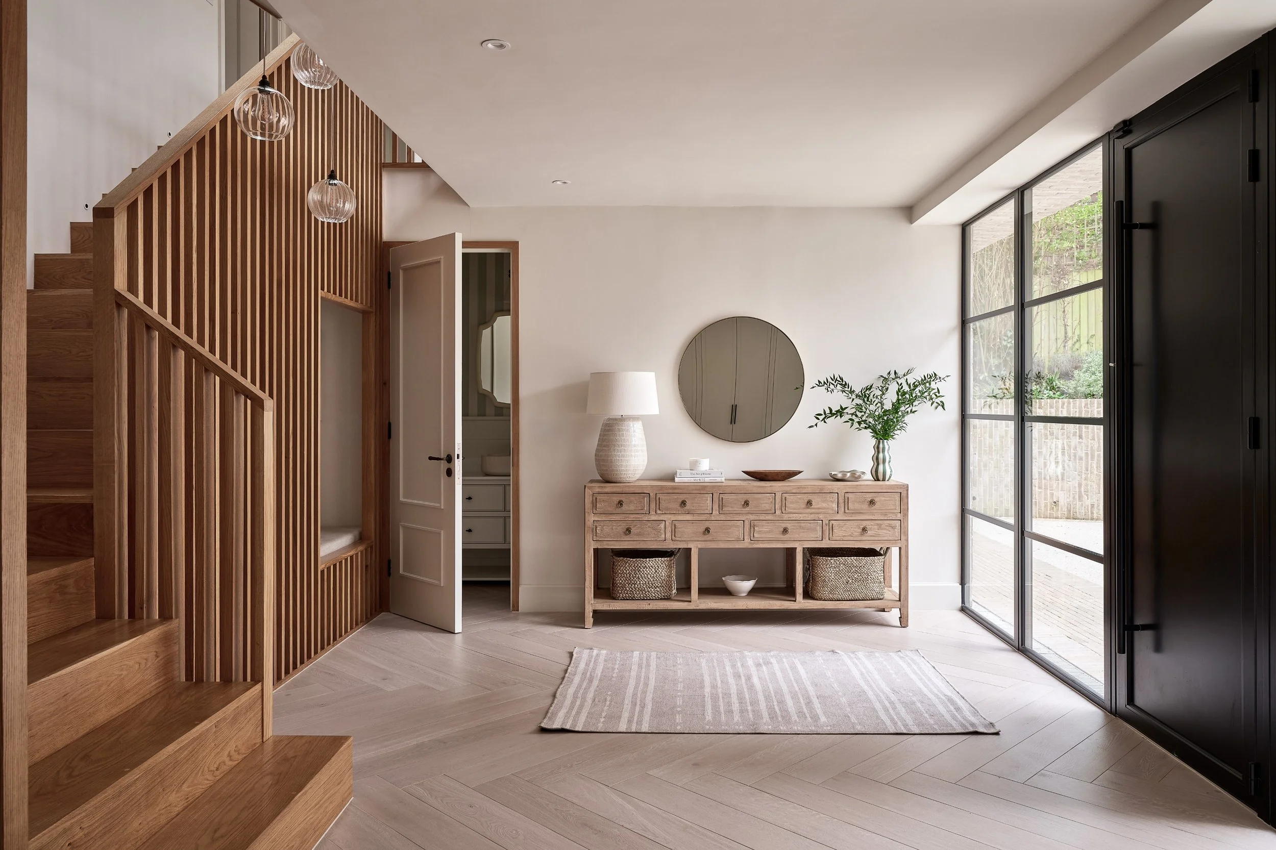 A modern living space with a wooden staircase to the left, a white door open to a closet or room, a wooden console table with decorative items, a round mirror on the wall, a table lamp, a potted plant, a small bowl, and baskets underneath. Large floo