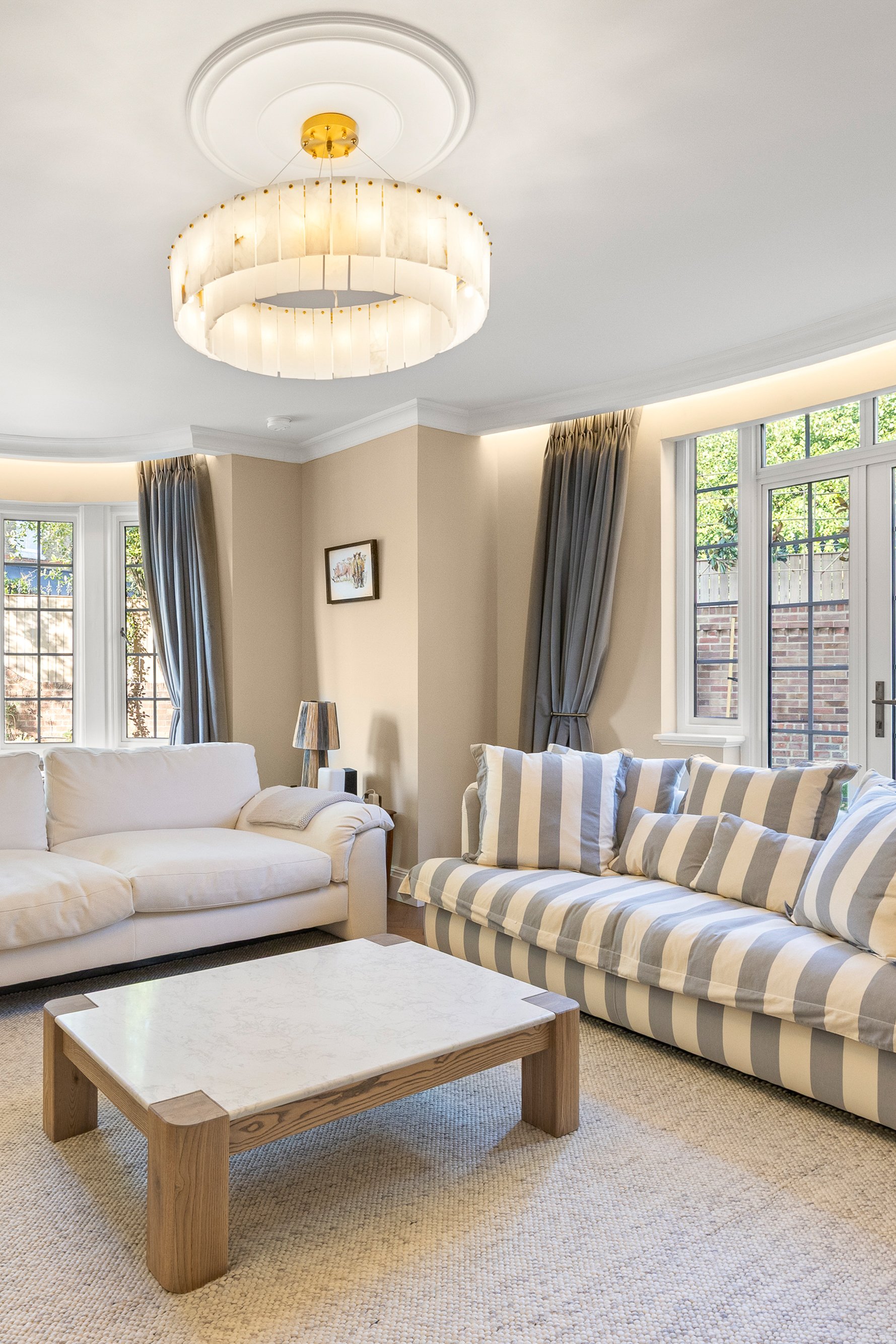 Living room with beige walls, two large bay windows with gray curtains, a white sofa, a striped gray and white armchair, a wooden coffee table with a marble top, and a ceiling chandelier with a circular design.