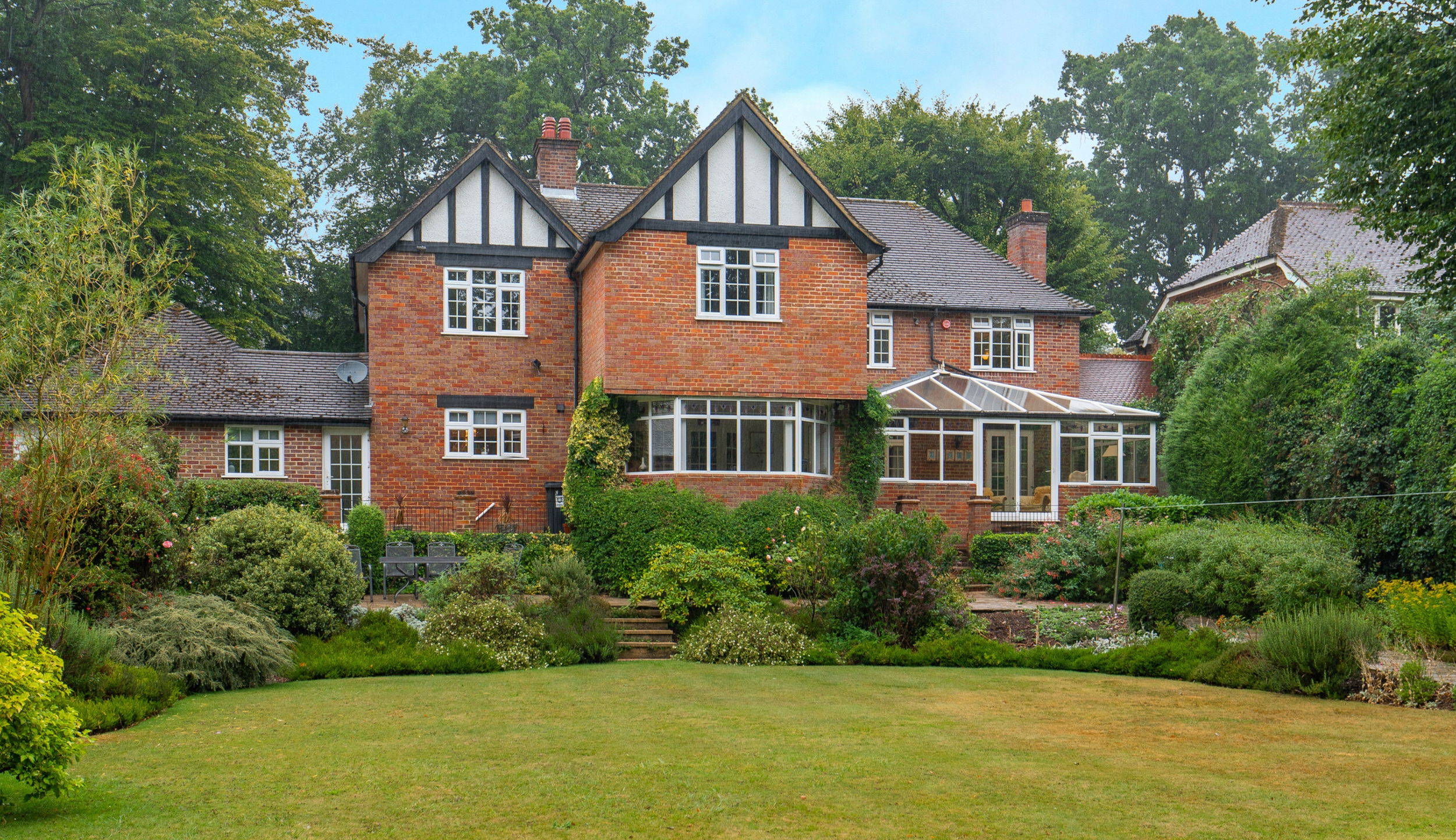 A large brick house with multiple stories, white-framed windows, and a glass-enclosed backyard porch. The house is surrounded by a well-maintained garden with grass, shrubs, and trees in the background.