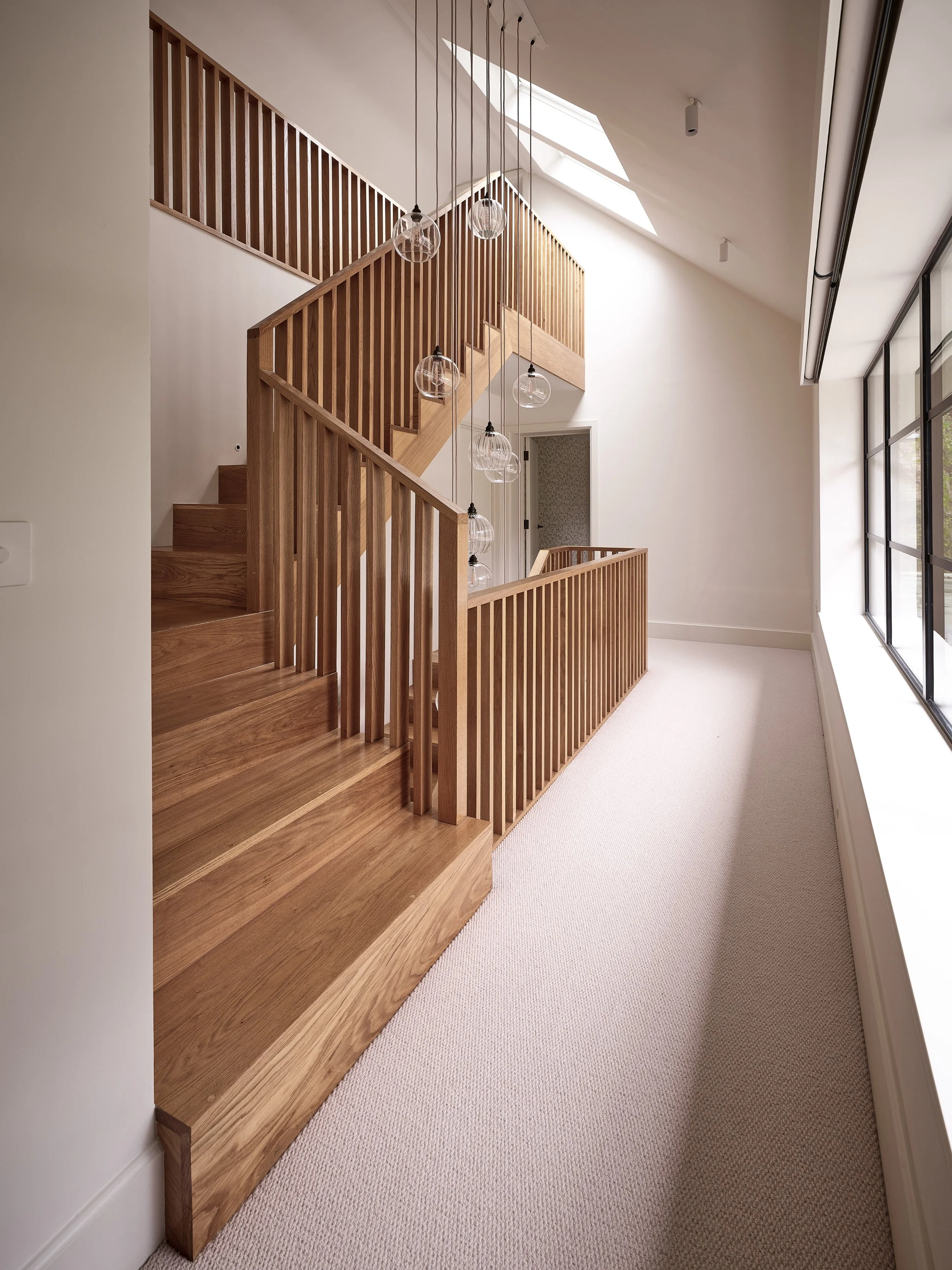 Interior view of a staircase with wooden railings and stairs, pendant lights hanging from the ceiling, large windows on the right, and skylights on the ceiling.