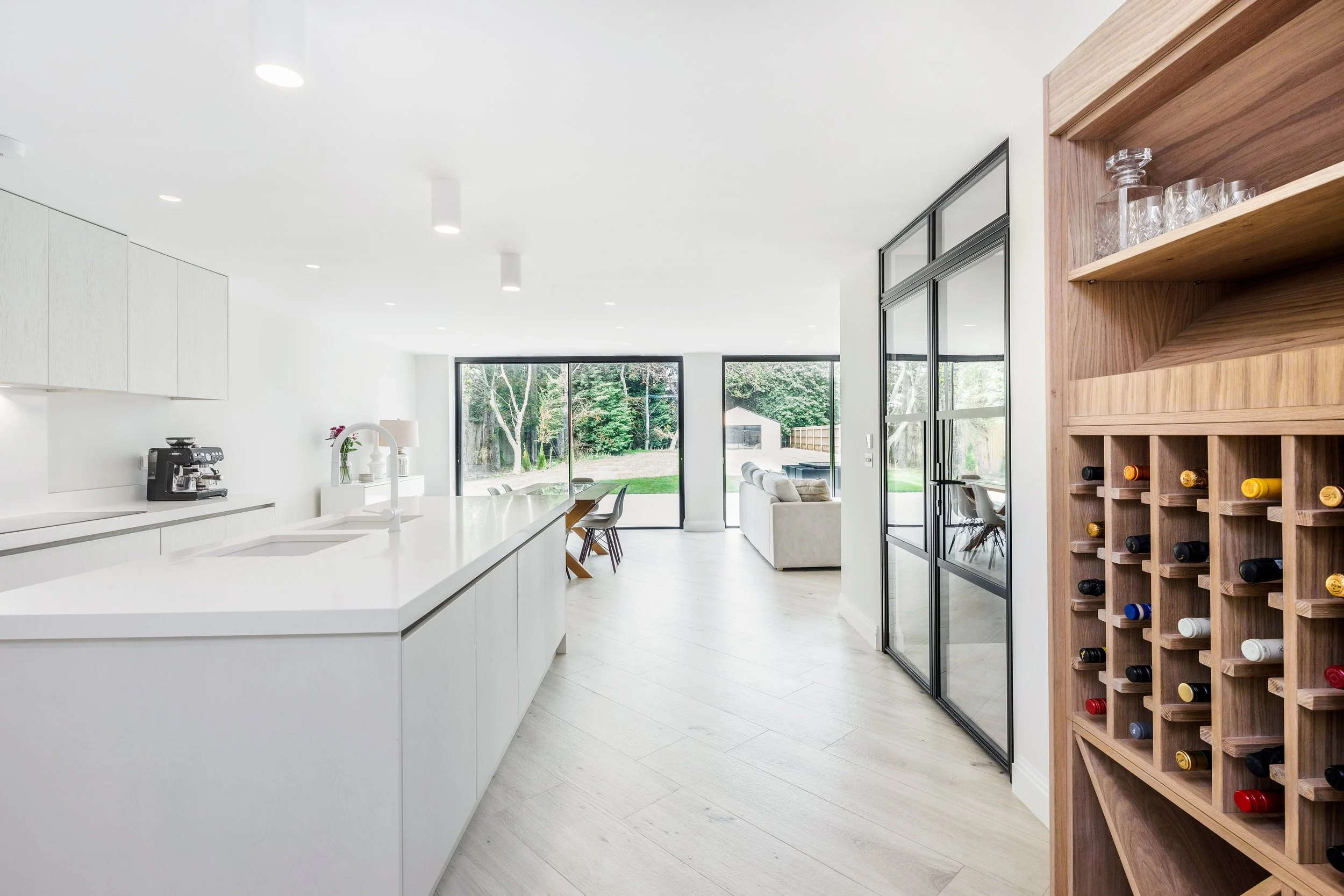 Modern open-concept kitchen and living room with large windows, white cabinets, a white island, a built-in wine rack, and a view of greenery outside.