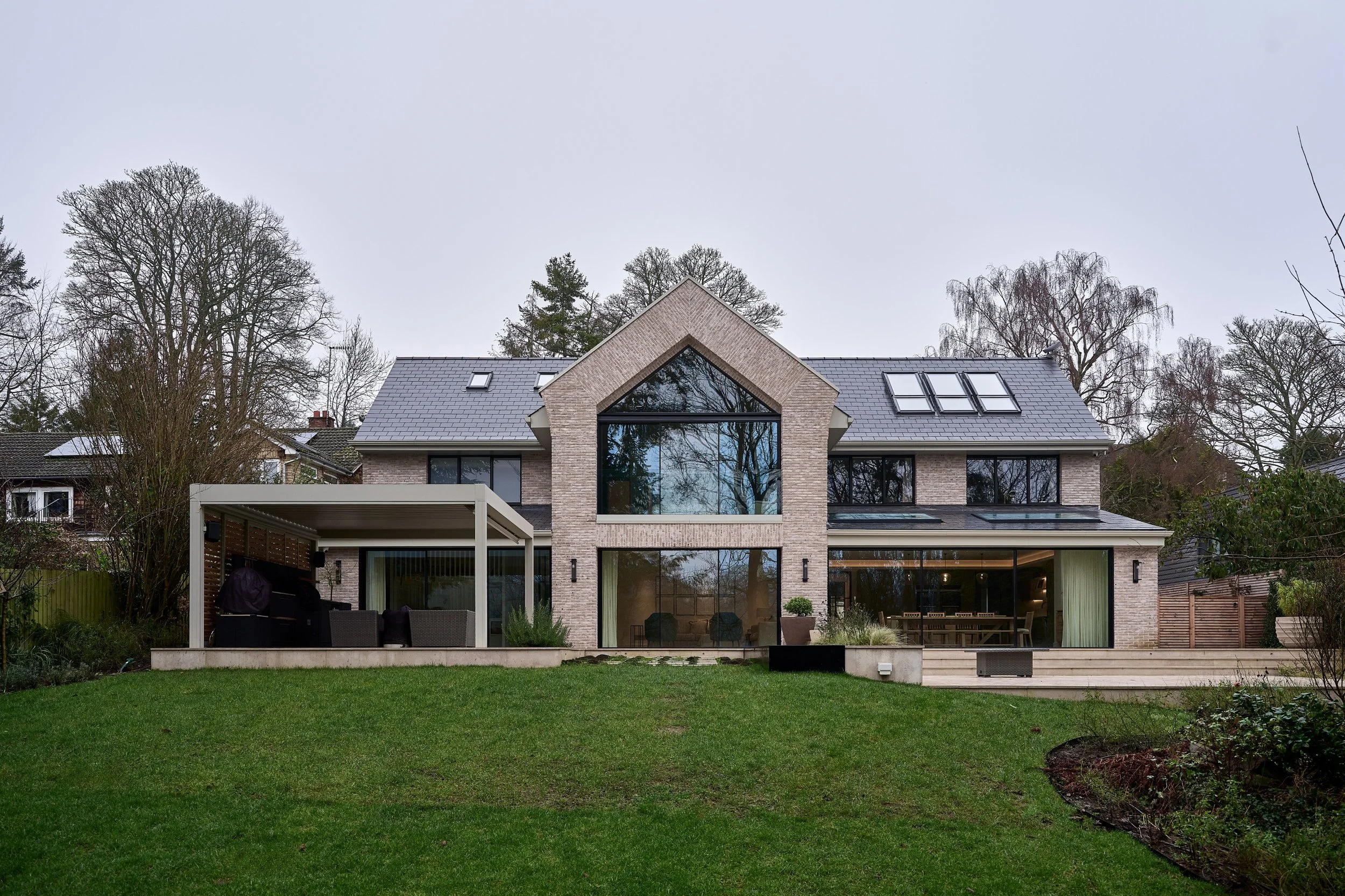 A modern two-story house with large windows and a sloped roof, surrounded by a lush lawn and tall trees on a cloudy day.