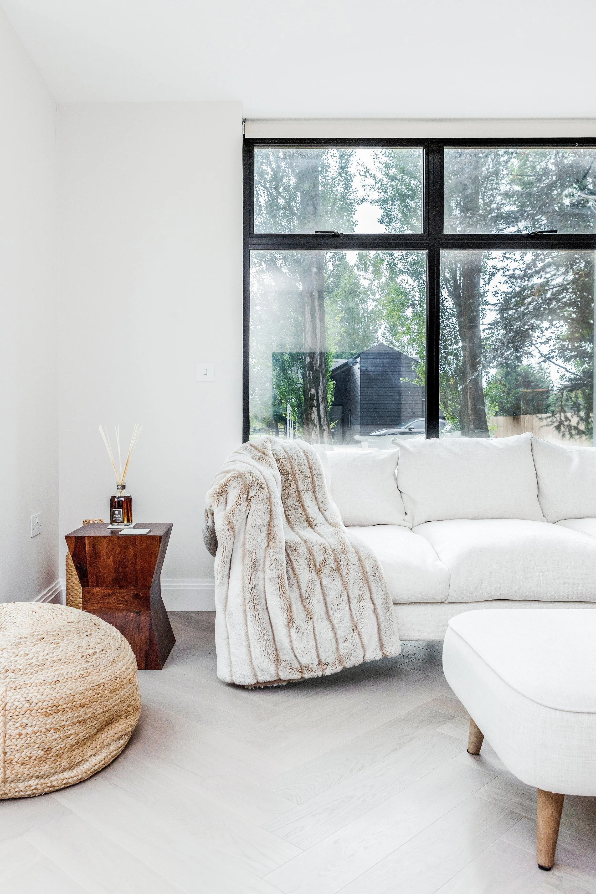 A modern living room with a large window, a white sofa with a plush fur throw, a wooden side table, a woven pouf, and a white chair.