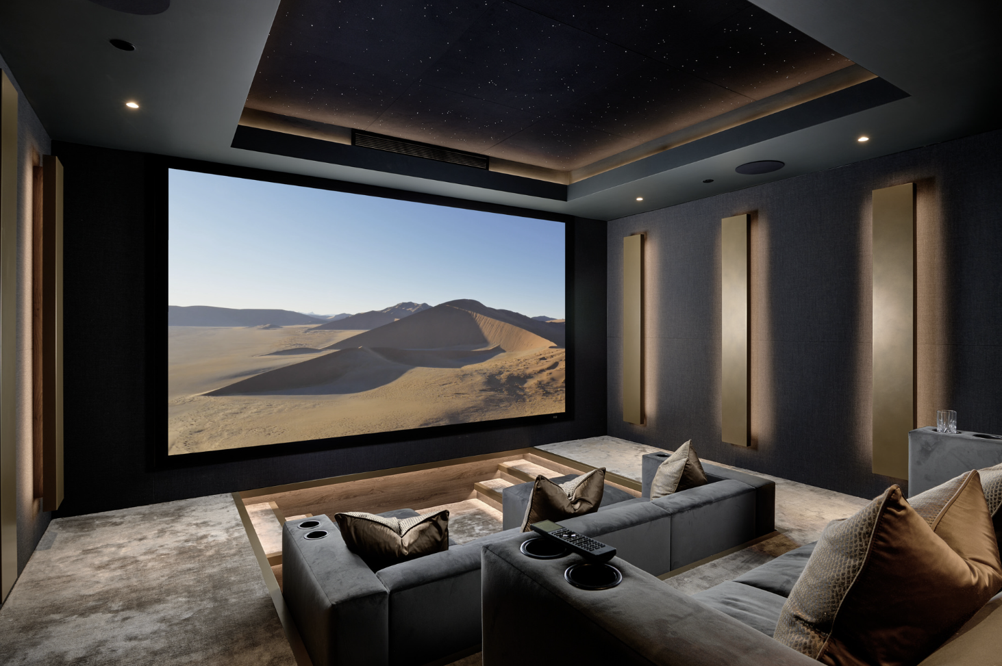 A home theater room with a large screen displaying a desert landscape with sand dunes. The room has dark walls, a plush gray sectional sofa with decorative pillows, built-in lighting, and a starry sky ceiling.