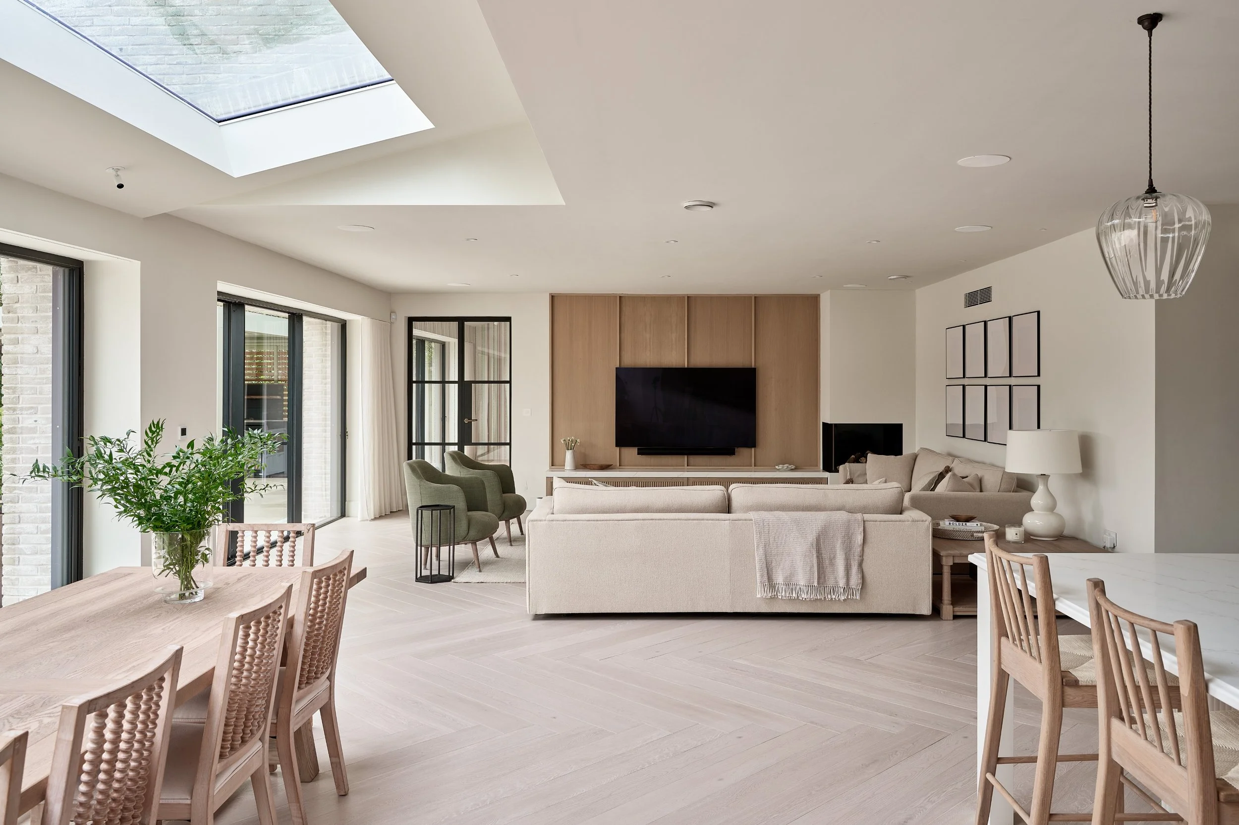 A modern living room with large windows and sliding glass doors, a ceiling with skylights, a beige sofa, green armchairs, a wall-mounted flat-screen TV, light wood flooring, and a dining table with wooden chairs.