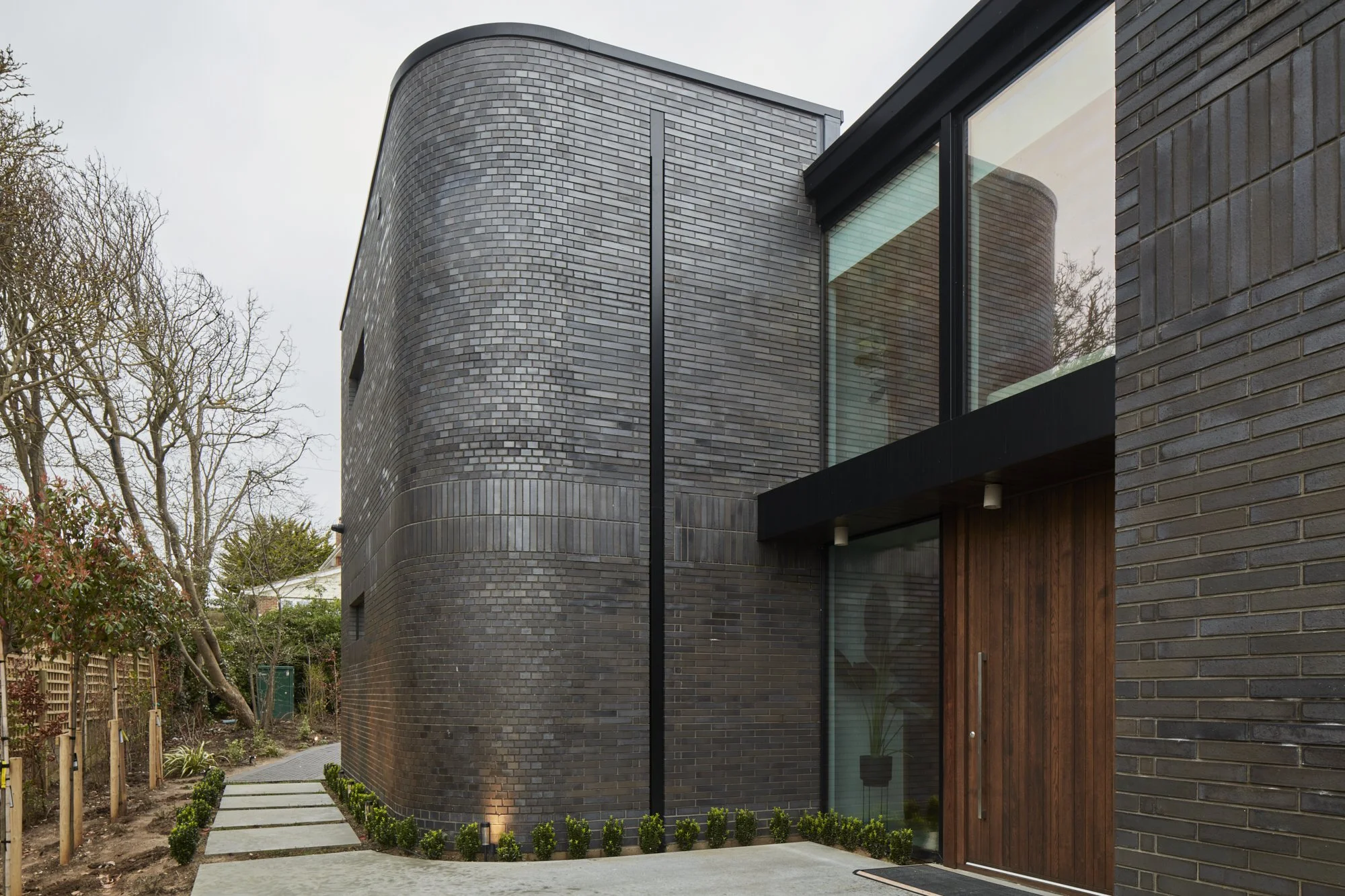 Modern house with curved black brick walls, a large glass window, wooden accents, and a pathway with small bushes leading to the entrance, surrounded by a garden and trees.