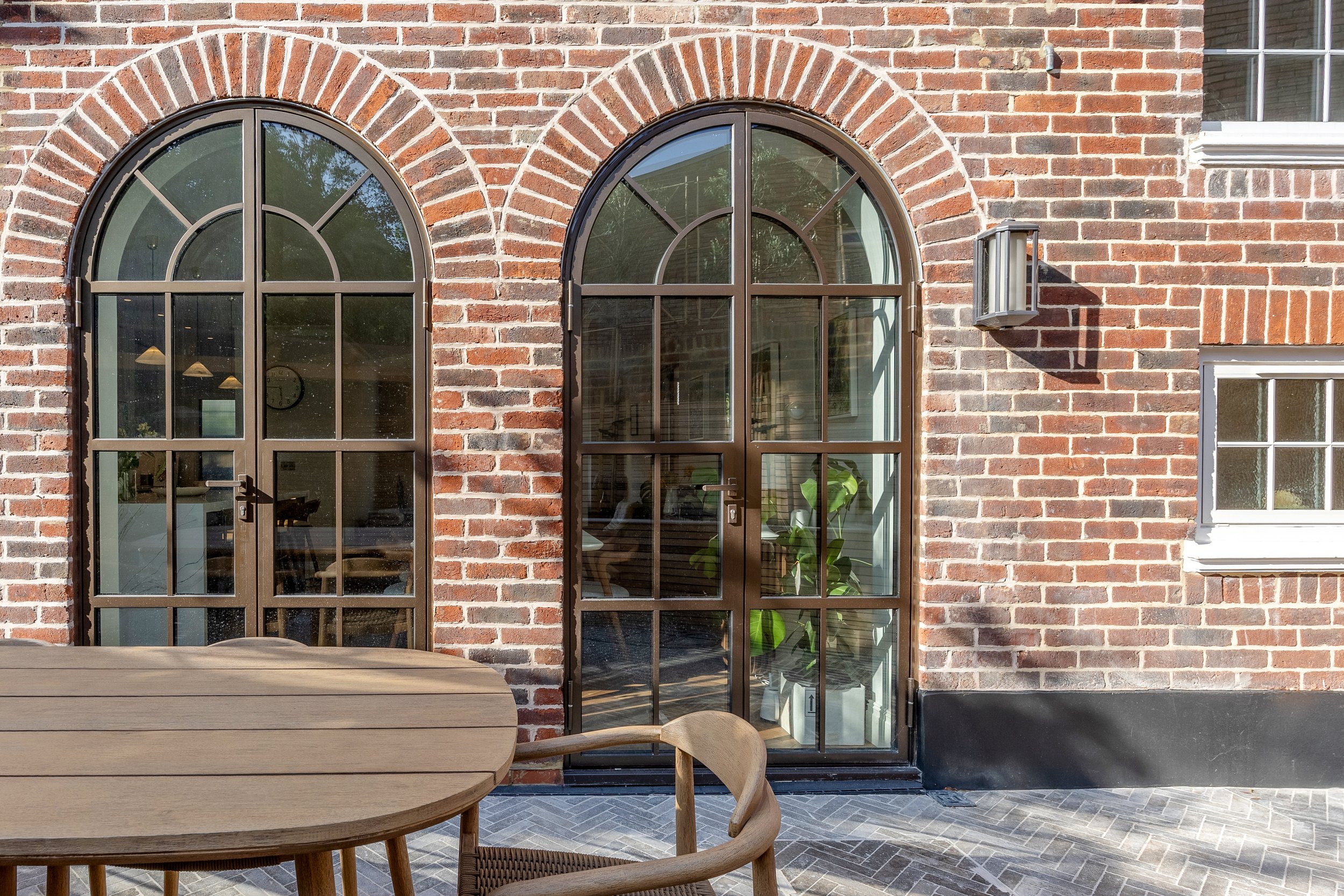 A brick exterior wall with three large arched windows, one door, a wall-mounted light fixture, and a portion of a white window frame. There is outdoor furniture including a rounded wooden table and chairs.