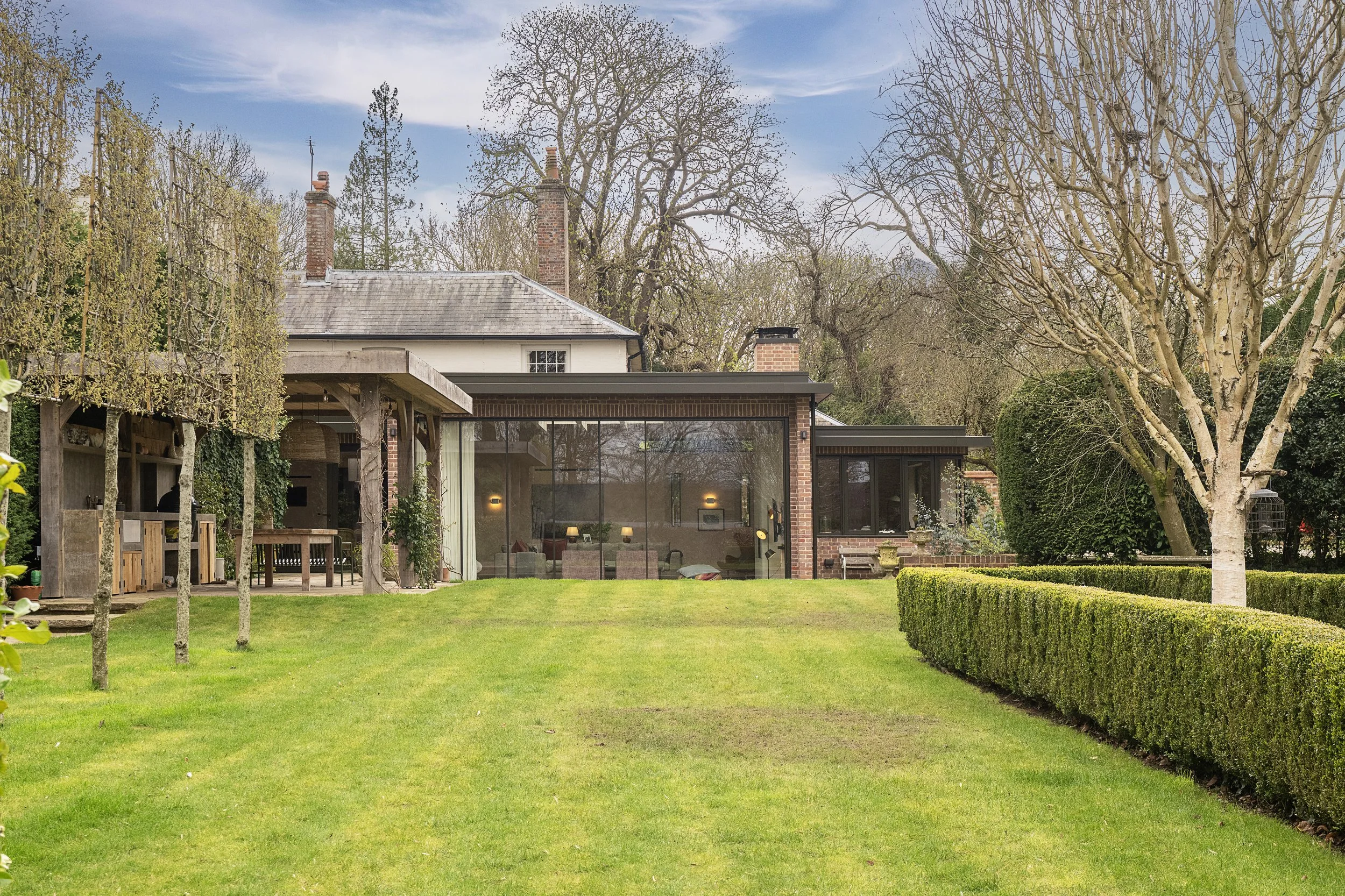 A backyard scene with a well-maintained lawn, a large leafless tree, and a modern house with large glass windows and brick accents. There is a covered patio area with outdoor furniture and a garden shed to the left.