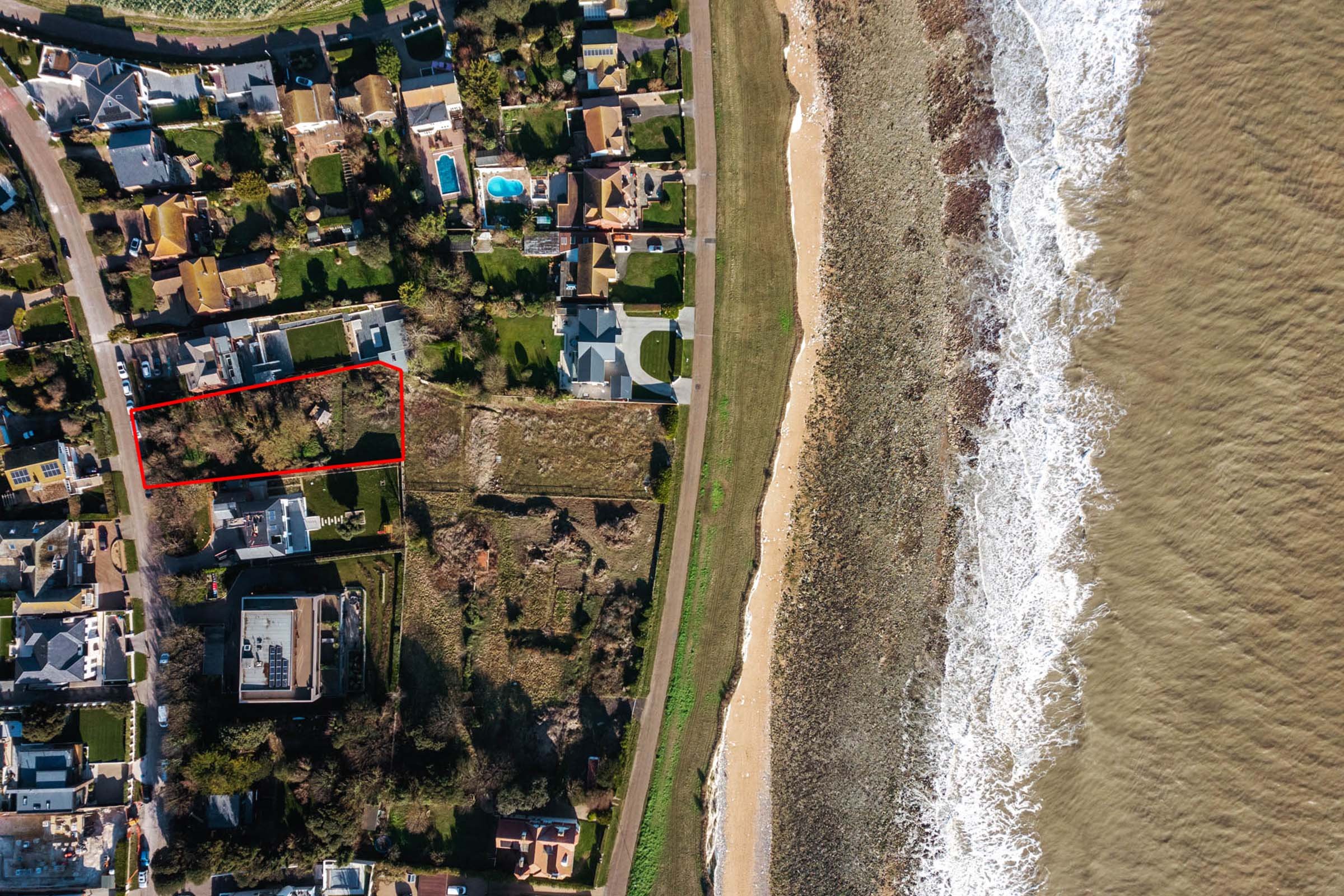 An aerial view of a coastal neighborhood with houses, trees, and backyard pools next to the beach and ocean.