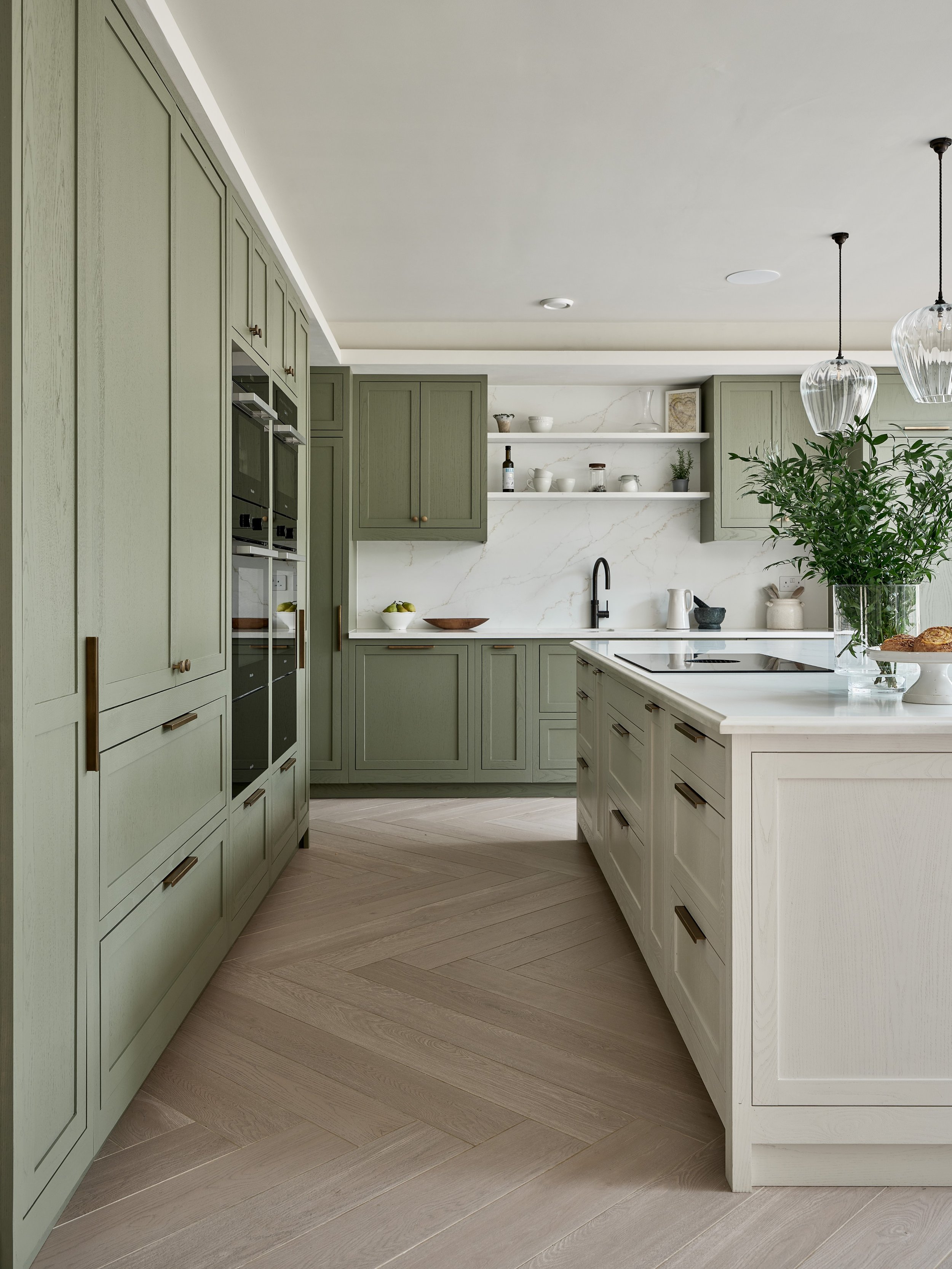 A modern kitchen with green and white cabinetry, open floating shelves with dishes and decor, a marble backsplash, and a large potted plant on a kitchen island.