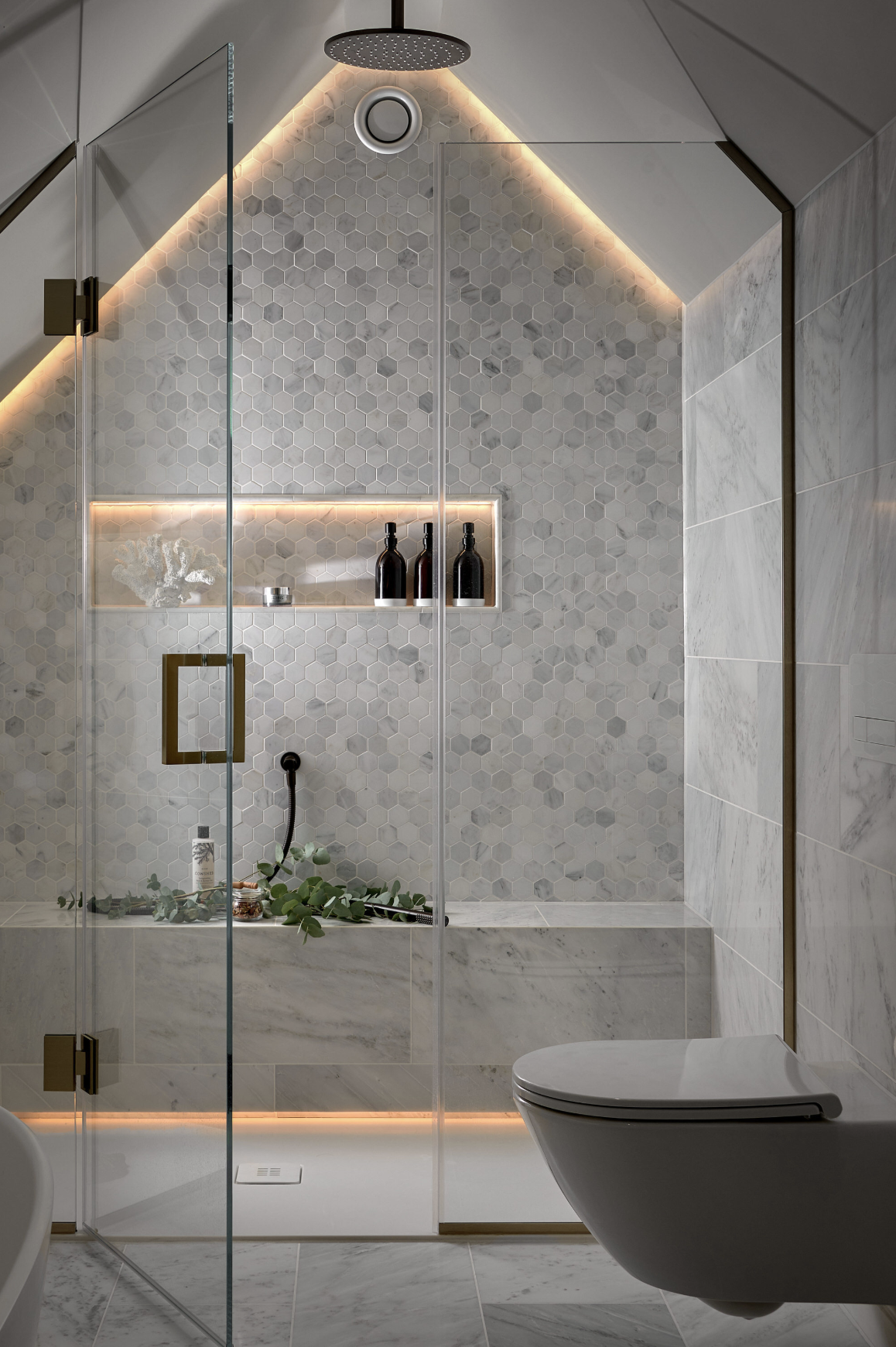 Modern bathroom with hexagonal gray tiles, a walk-in shower with glass doors, a round ceiling showerhead, built-in shelf with bottles and decor, and a white toilet.