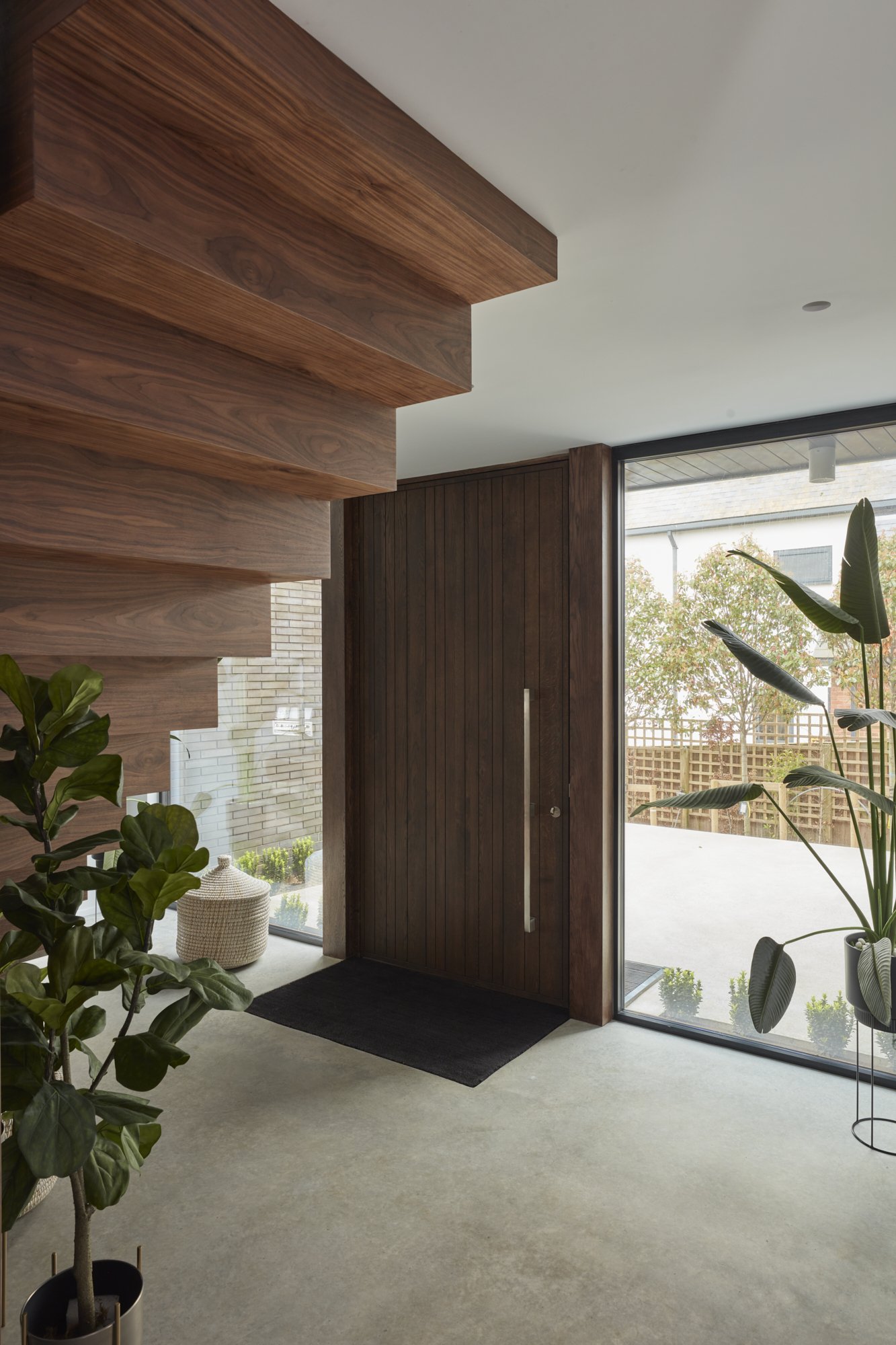 Interior view of a modern living space with a wooden staircase, large floor-to-ceiling windows, potted plants, and a black doormat near a dark wooden door.