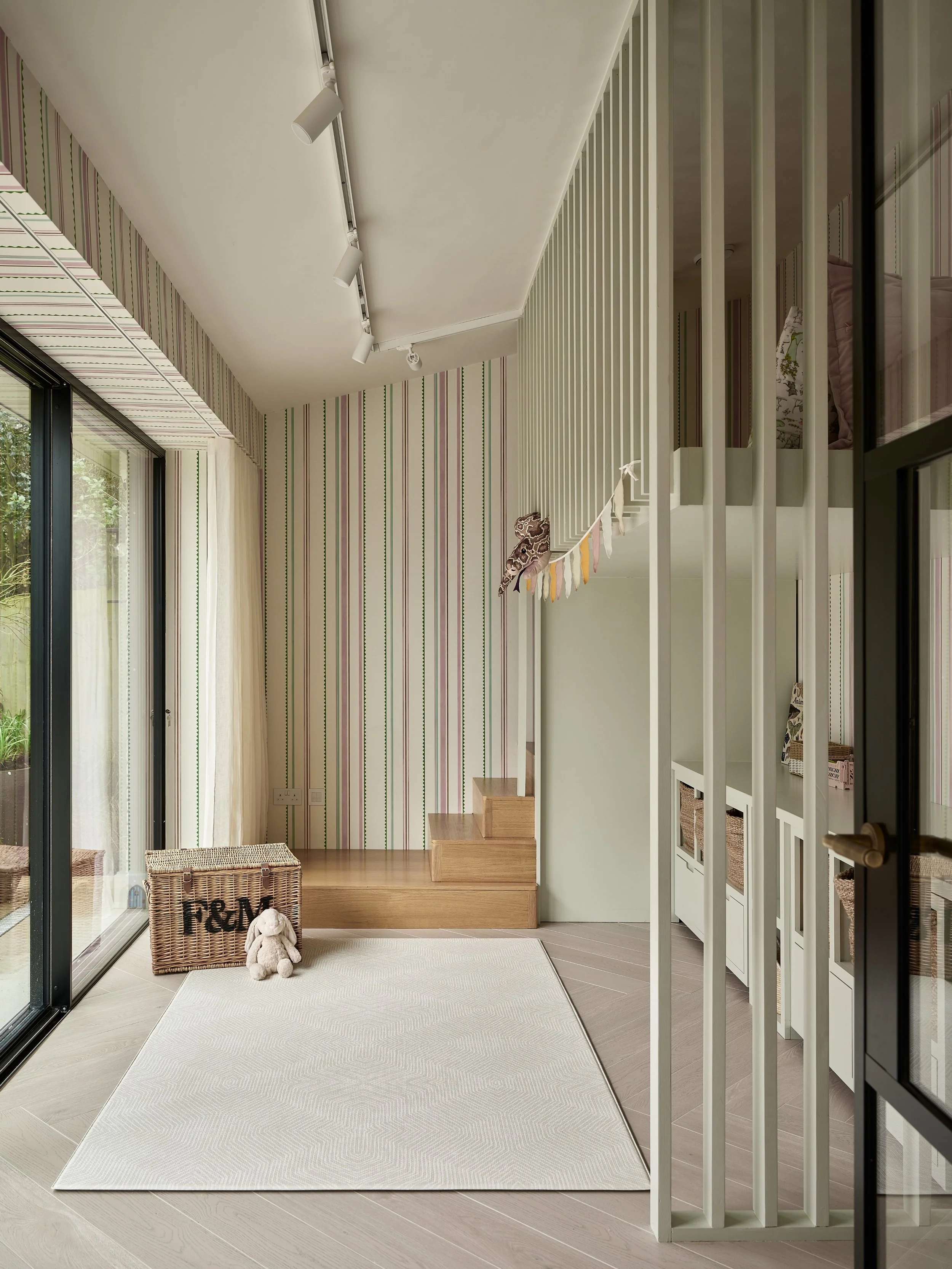 Interior view of a modern home entrance area with wooden steps leading up, a white rug on the floor, corner with a wicker basket, plush toy, and a cloth bag. A large sliding glass door opens to the outside with trees visible. The wall has vertical st