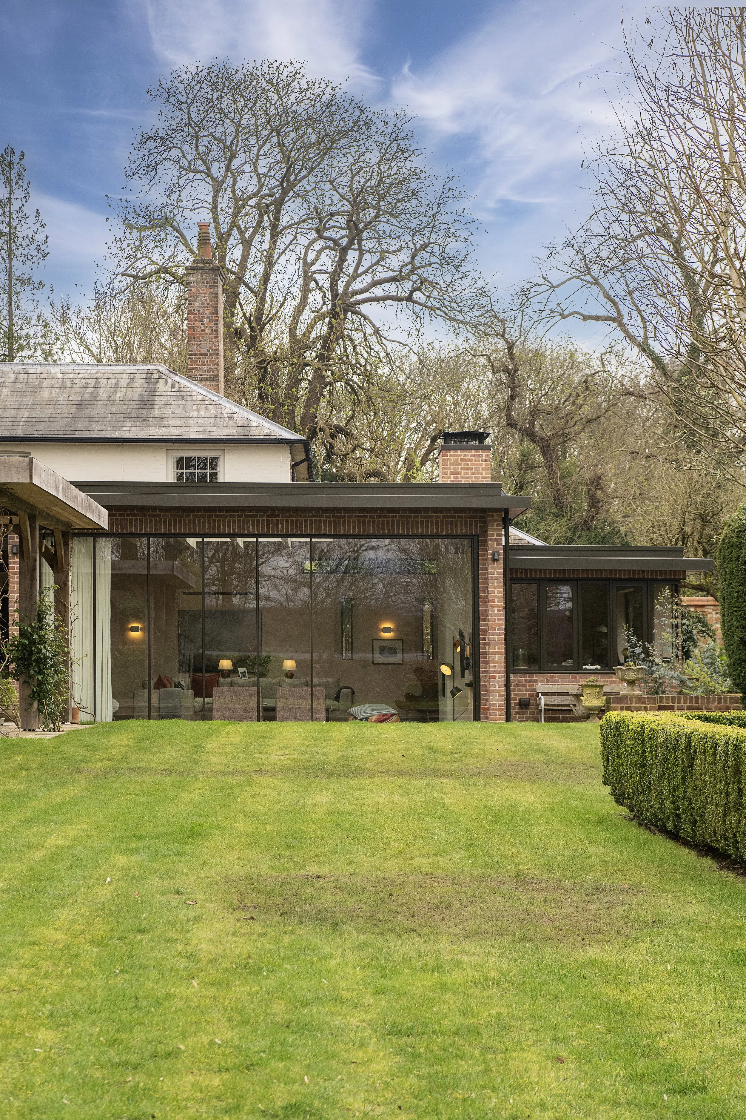View of a modern house with large glass windows, a manicured lawn, bushes, and tall trees with no leaves under a partly cloudy sky.