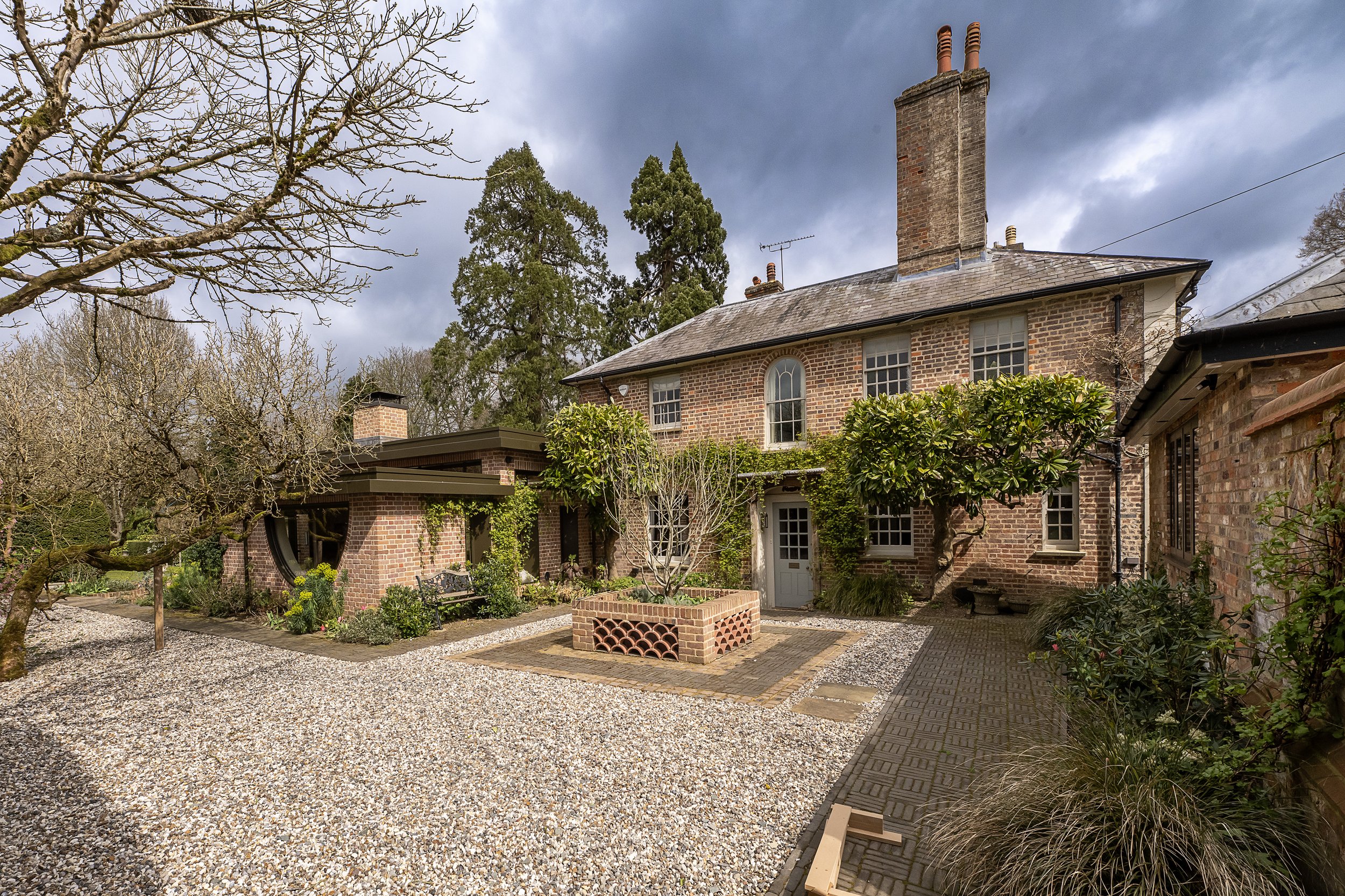A brick house with a chimney, multiple windows, and a garden with trees and bushes. The sky is cloudy.