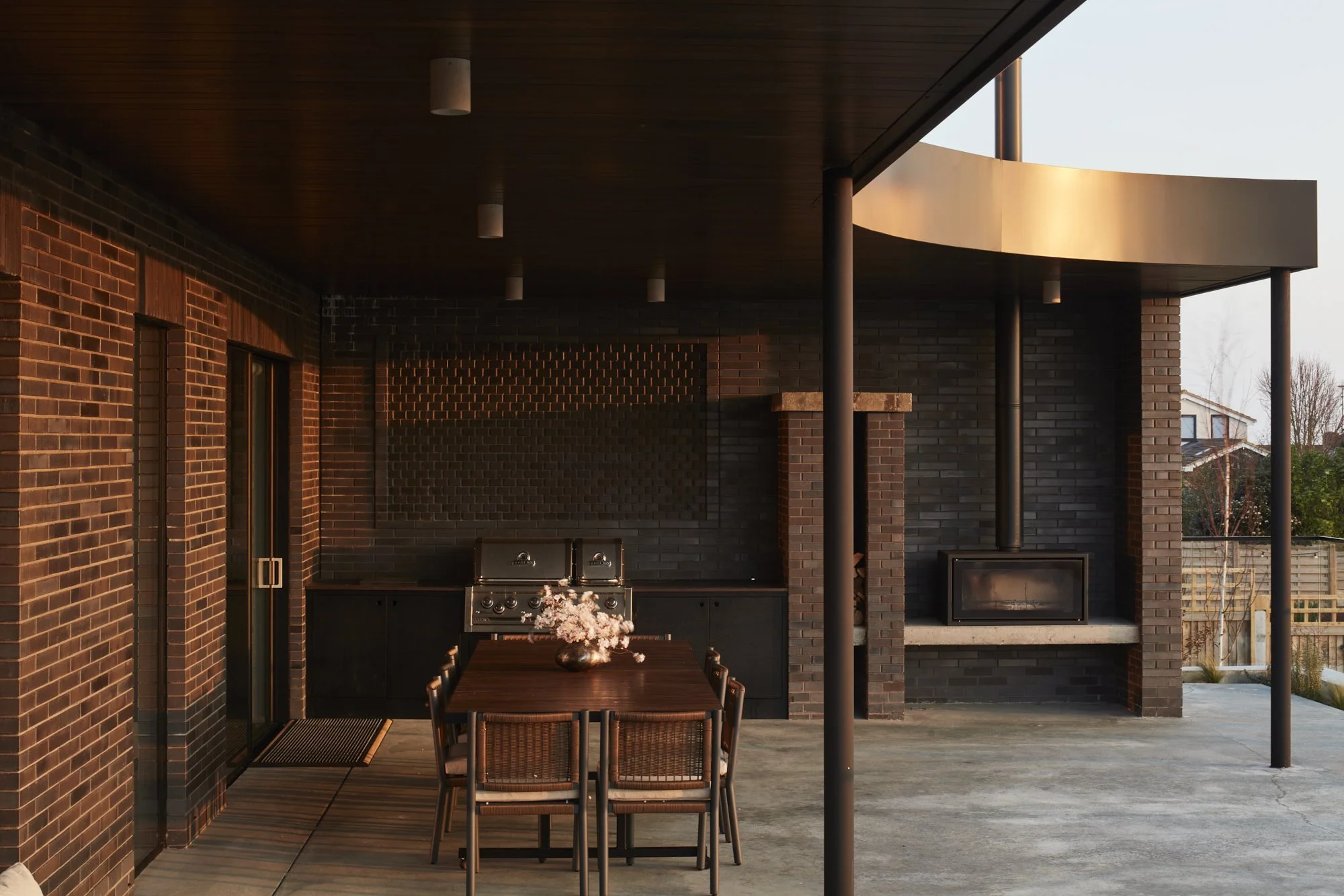 Outdoor patio area with brick walls, wooden ceiling, a dining table with six chairs, a fireplace, and a barbecue grill, illuminated by warm sunlight during sunset.