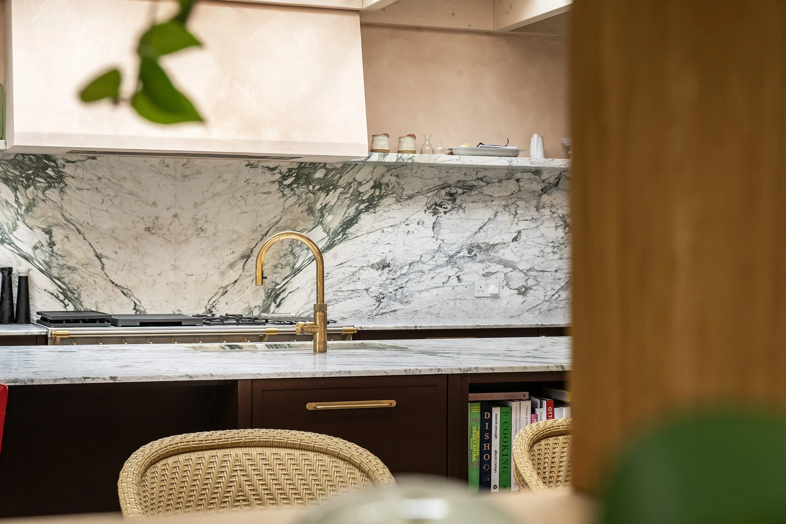 A kitchen with a marble countertop and backsplash, a gold faucet, a stove, and kitchen books on a shelf below the counter. There are chairs with woven backs in the foreground, and blurred green leaves in the upper left corner.