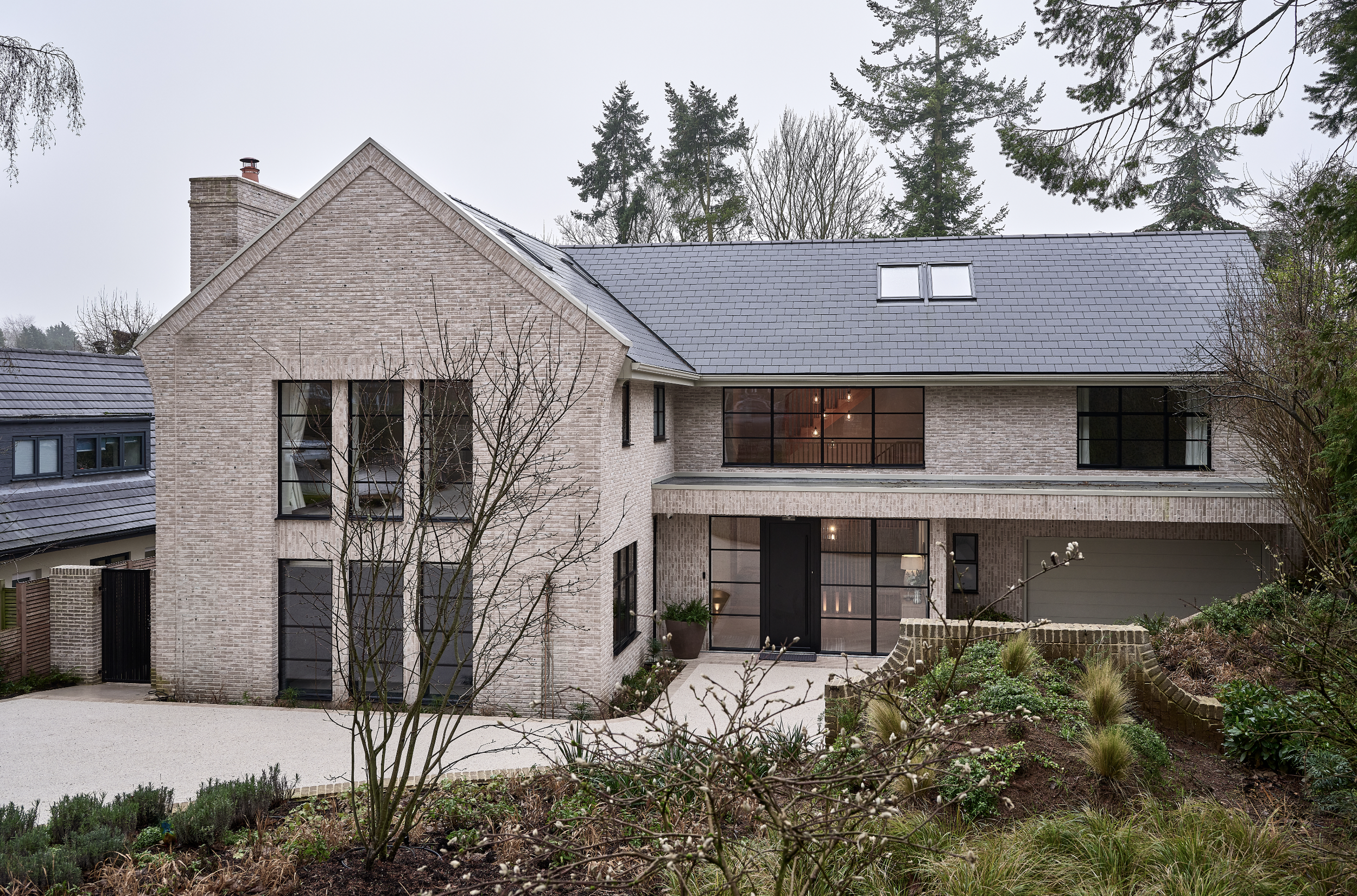 Modern multi-story house with brick exterior, large windows, and a sloped roof in a garden setting with trees and plants.